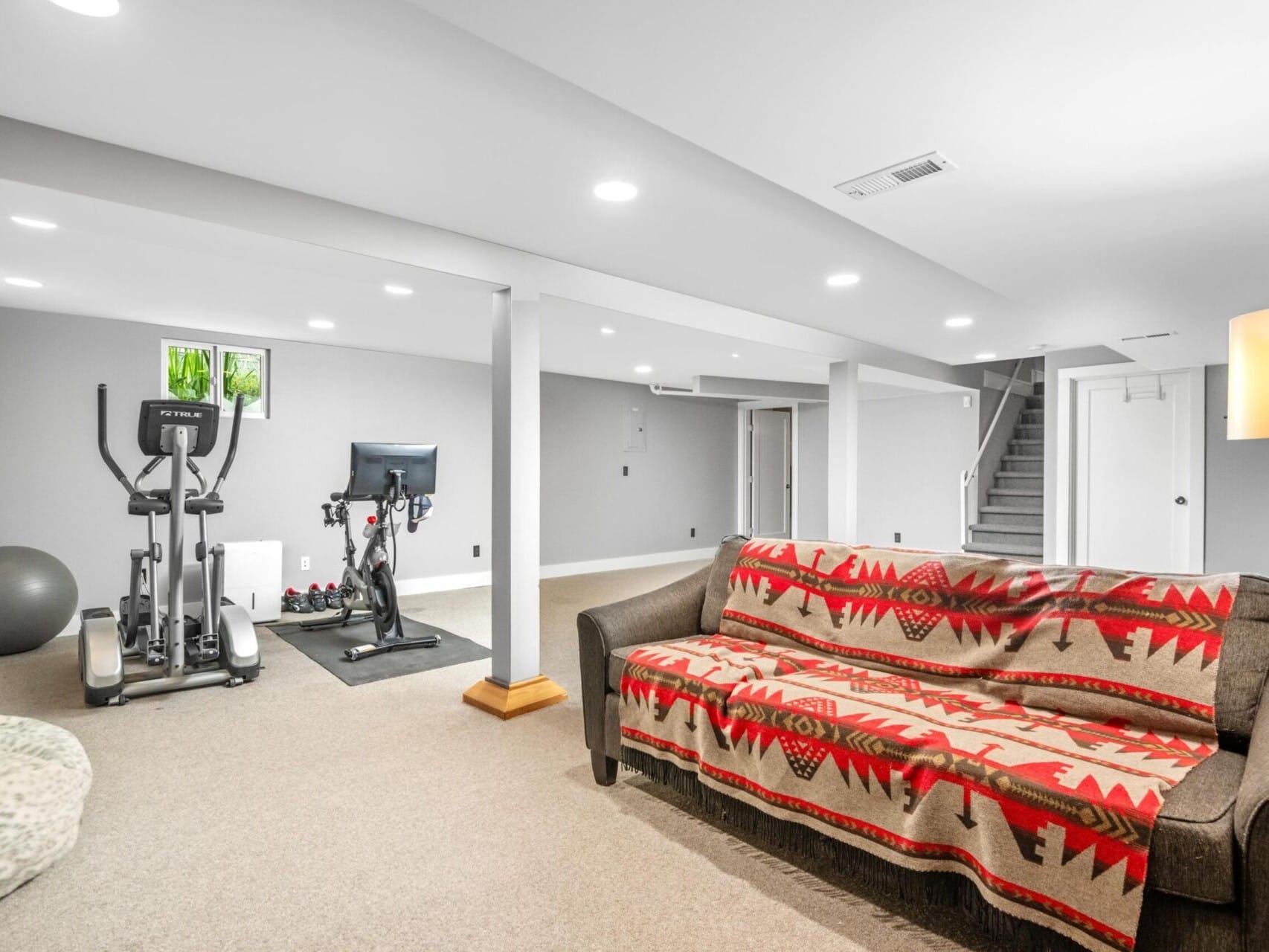 A basement room in Portland Oregon Real Estate features exercise equipment, including a stationary bike and an elliptical machine. A gray couch with a red and gray patterned blanket is in the foreground, all illuminated by bright recessed lighting.