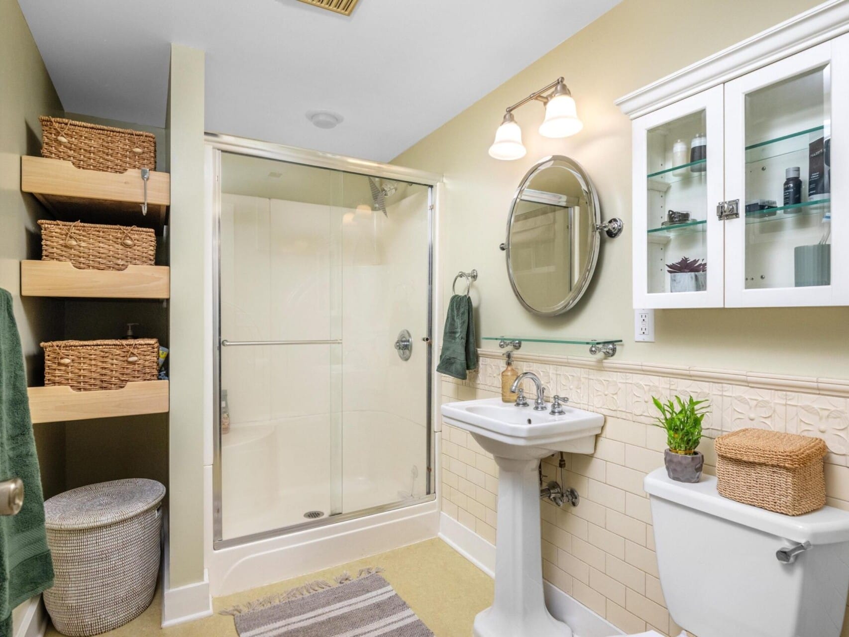 A bathroom with beige walls features a glass shower, a pedestal sink, and a toilet. A round mirror and wall cabinet hang above the sink, with shelves holding wicker baskets. Recommended by a top Portland Oregon realtor, this serene space includes a towel rack and a plant on the toilet tank.