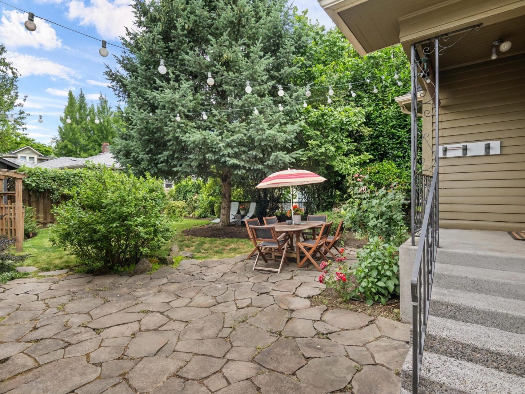 A backyard patio featuring a stone surface, outdoor dining set with wooden chairs, and a striped umbrella offers the perfect Portland Oregon real estate charm. String lights hang above, while lush greenery surrounds the space. Steps lead to the house on the right.