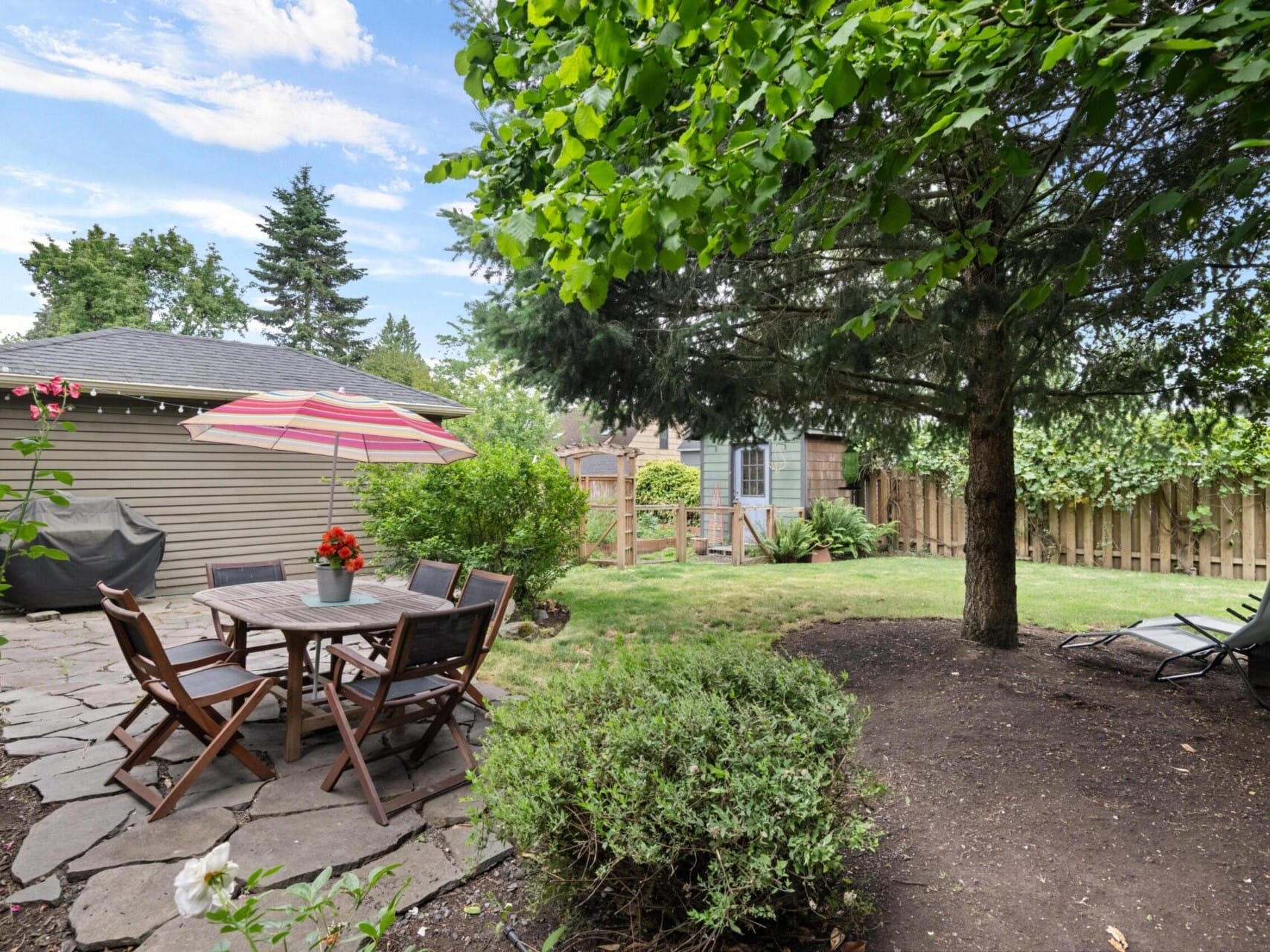 A backyard oasis perfect for entertaining, featuring a stone patio with a wooden table and chairs under an umbrella. Nearby, the Portland Oregon real estate includes a grill, lush lawn, trees, and two lounge chairs on the grass next to a small shed and wooden fence.