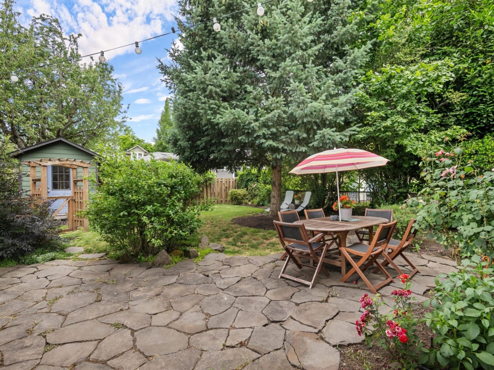 A serene backyard with a stone patio, wooden dining set under an umbrella, and lush greenery, exemplifies Portland Oregon real estate charm. A small shed is visible in the background. String lights hang above, adding a cozy touch to this inviting garden setting.