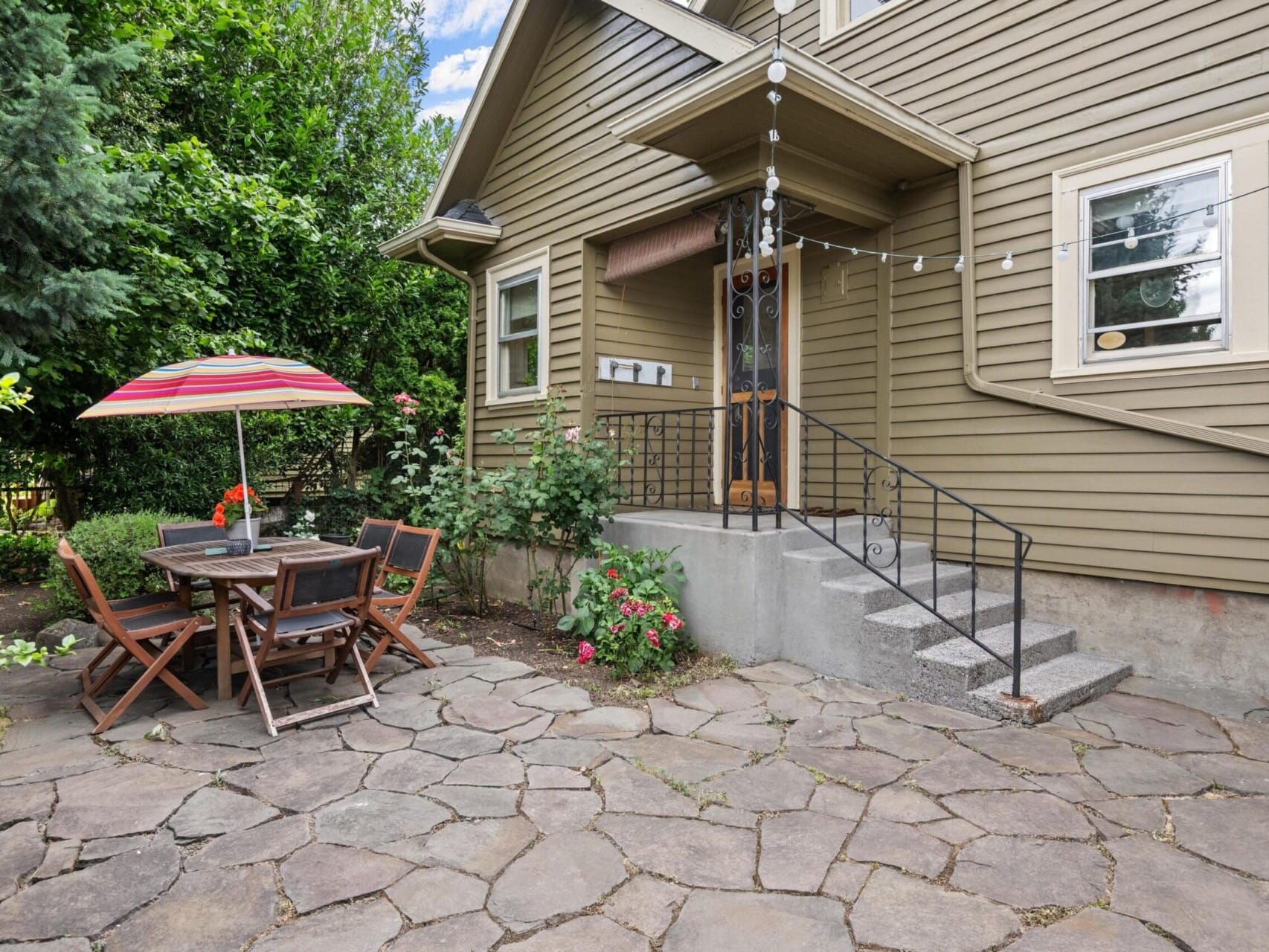 A lovely patio with wooden furniture, including a table and chairs, sits under a colorful striped umbrella. Surrounded by greenery and flowers, this Portland Oregon real estate gem features a house with brown siding, steps leading up to the door, and stone paving that covers the ground.