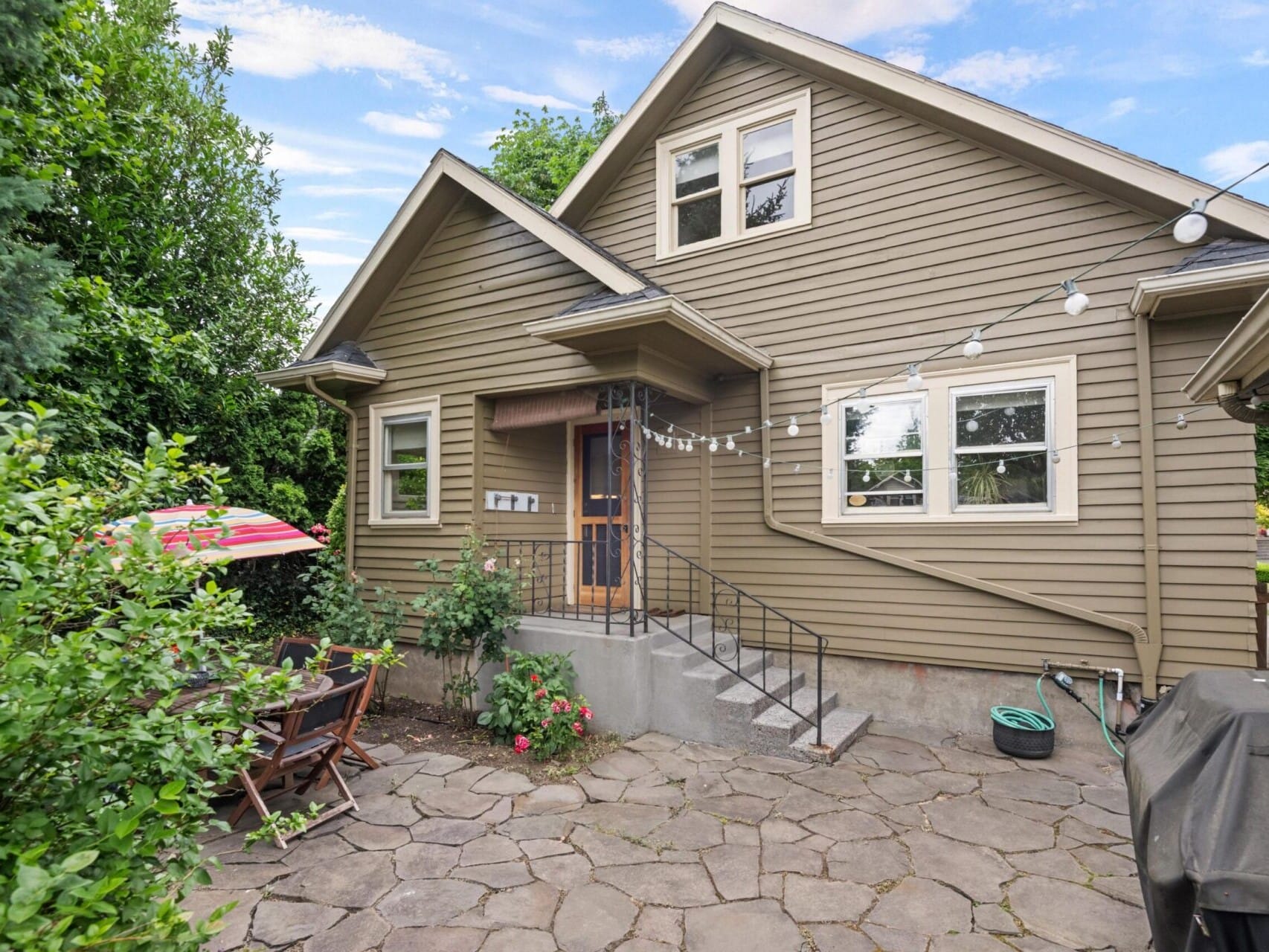 A quaint house with a stone patio, surrounded by greenery, embodies Portland Oregon real estate charm. The patio features a wooden table with an umbrella and string lights overhead. The house has a gabled roof and several windows, creating a cozy outdoor space.