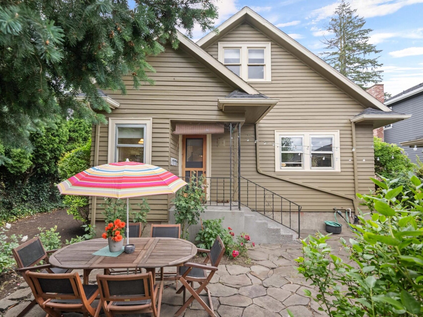 A cozy house with beige siding and a small porch sits invitingly in the backdrop of Portland, Oregon real estate. In the foreground, there's a patio with a round wooden table, colorful umbrella, and chairs amidst lush greenery. The scene is completed by a clear blue sky overhead.