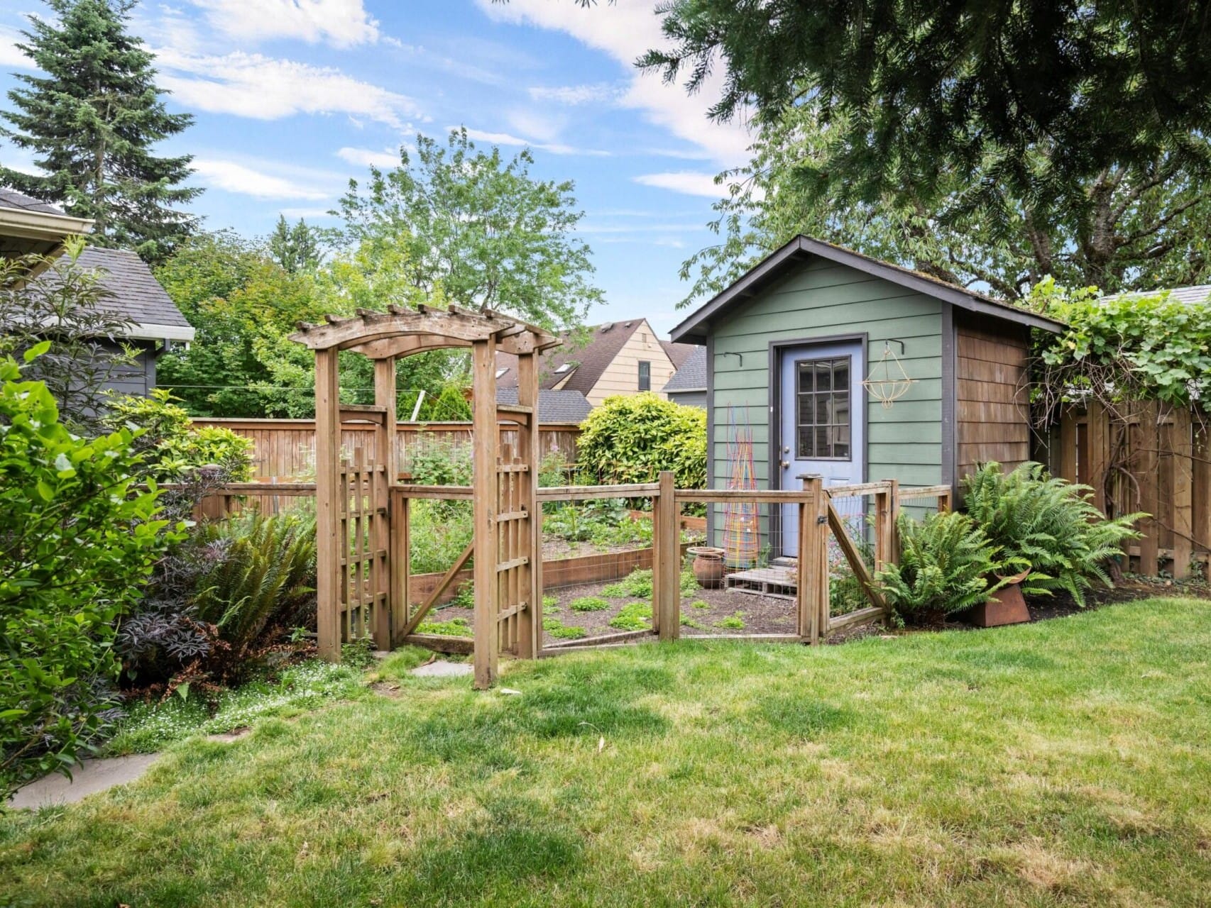 A lush backyard garden with a small green shed, a wooden fence, and an arched gate. The yard, perfect for any Portland Realtor listing, features vibrant grass, ferns, and various plants amid trees and neighboring homes under a partly cloudy sky.