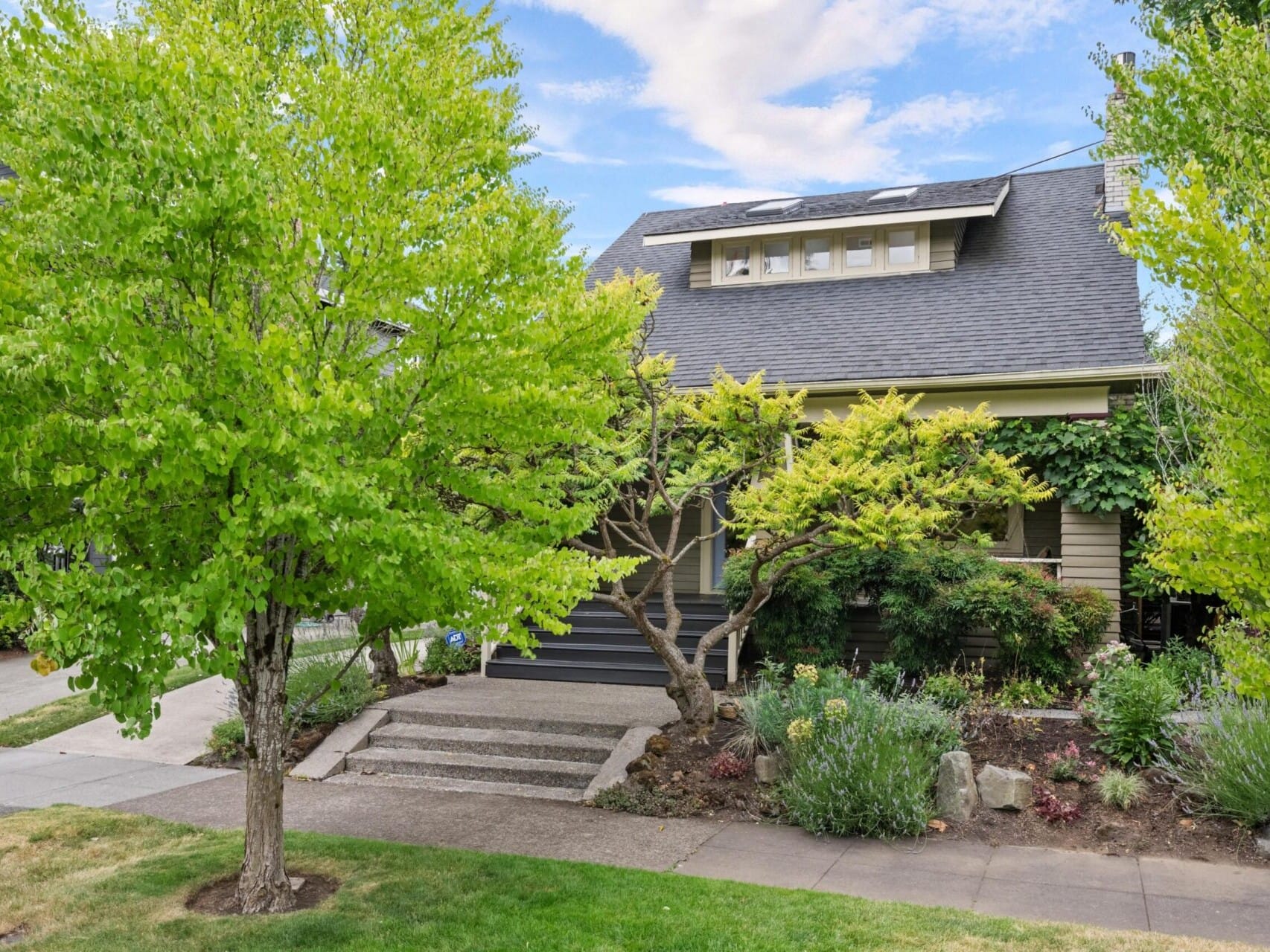 A two-story house with a dark roof and beige exterior, partially obscured by lush green trees and shrubs, showcases the charm of Portland Oregon real estate. A concrete walkway leads to the front steps, surrounded by landscaped greenery under a partly cloudy sky.