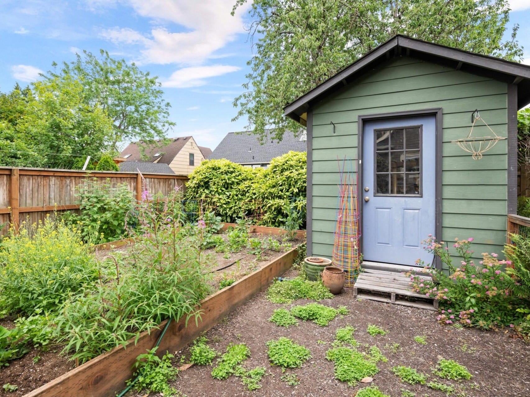 A charming garden scene with a green shed featuring a blue door graces this Portland Oregon real estate listing. Raised garden beds, bursting with various plants and flowers, sit beside it. The area is enclosed by a wooden fence, embraced by trees and a partly cloudy sky overhead.