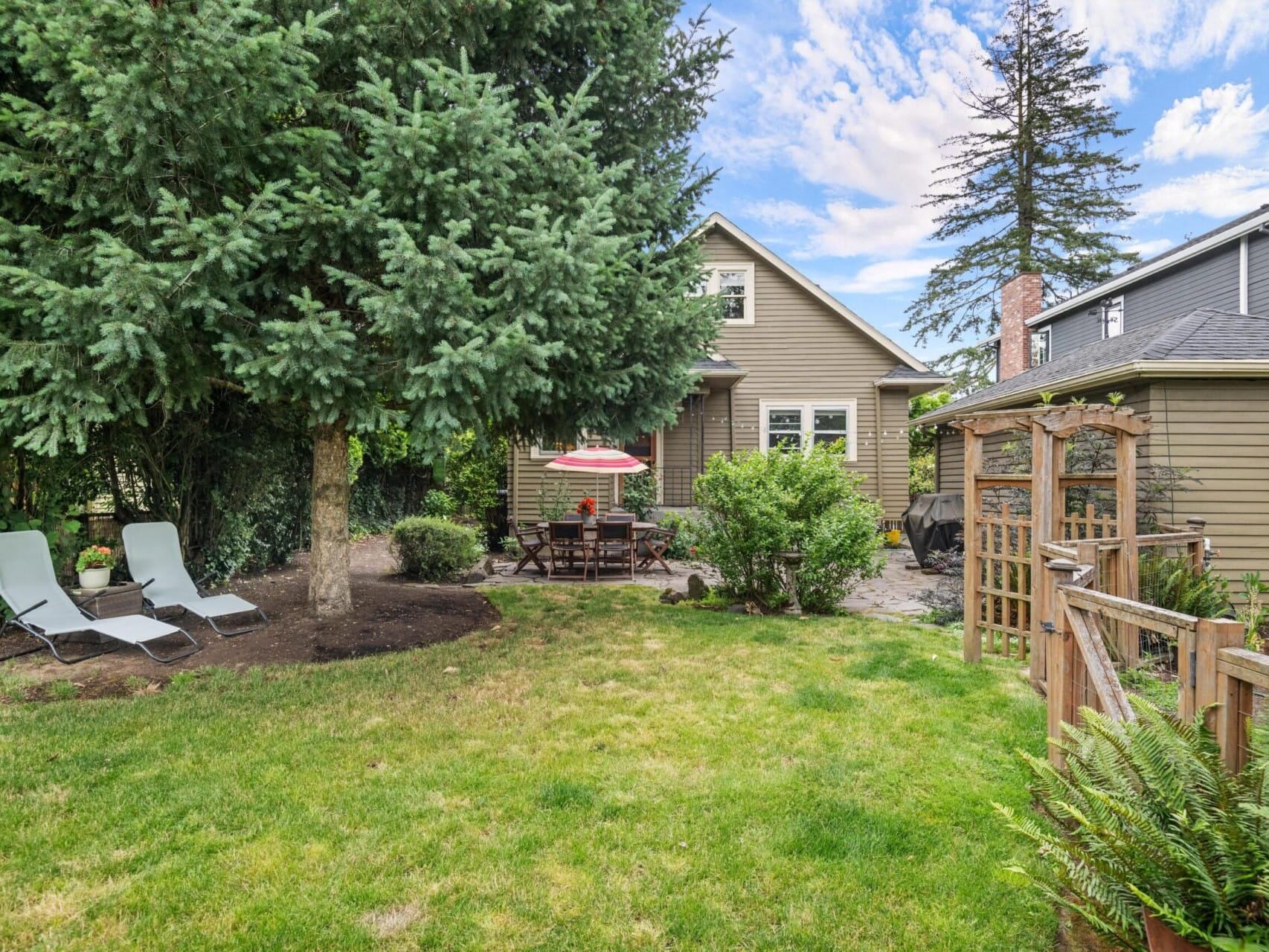 A quaint backyard showcases a cozy house with a sloped roof, reminiscent of Portland Oregon real estate charm. Two lounge chairs rest under a large tree, while a dining set with a red umbrella sits nearby, embraced by lush greenery and a wooden fence. The sky adds character with its partly cloudy canvas.