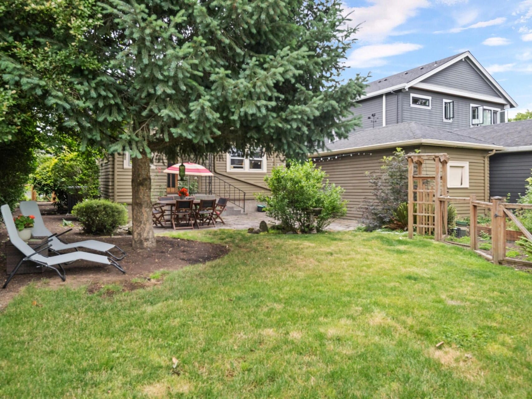 A backyard with green grass and trees showcases two lounge chairs to the left and a patio set with a red umbrella near a house in the background. This peaceful, well-maintained oasis is perfect for relaxation and highlights why Portland Oregon real estate is so desirable.