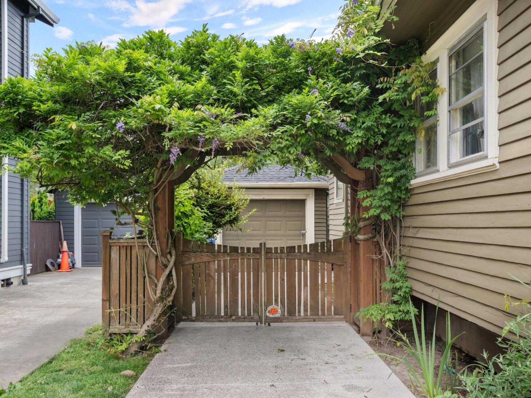 A charming wooden gate covered with lush green vines and purple flowers creates a shaded archway, welcoming you to this Portland Oregon real estate gem. The gate opens to a path leading to a garage, with houses on either side under a partly cloudy sky.