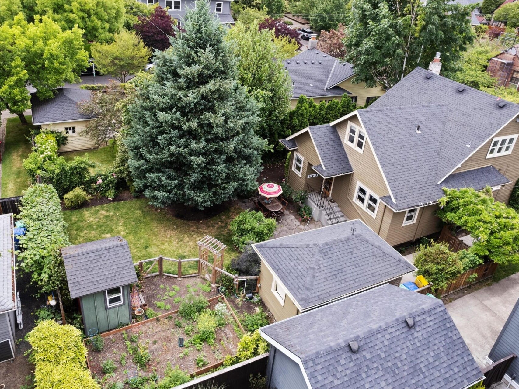 Aerial view of a Portland Oregon real estate gem: a beige house with a dark roof, nestled in greenery. It boasts a vegetable garden, large tree, and patio with an umbrella. Nearby are additional small buildings and parked cars, making it an ideal showcase for any top real estate agent.