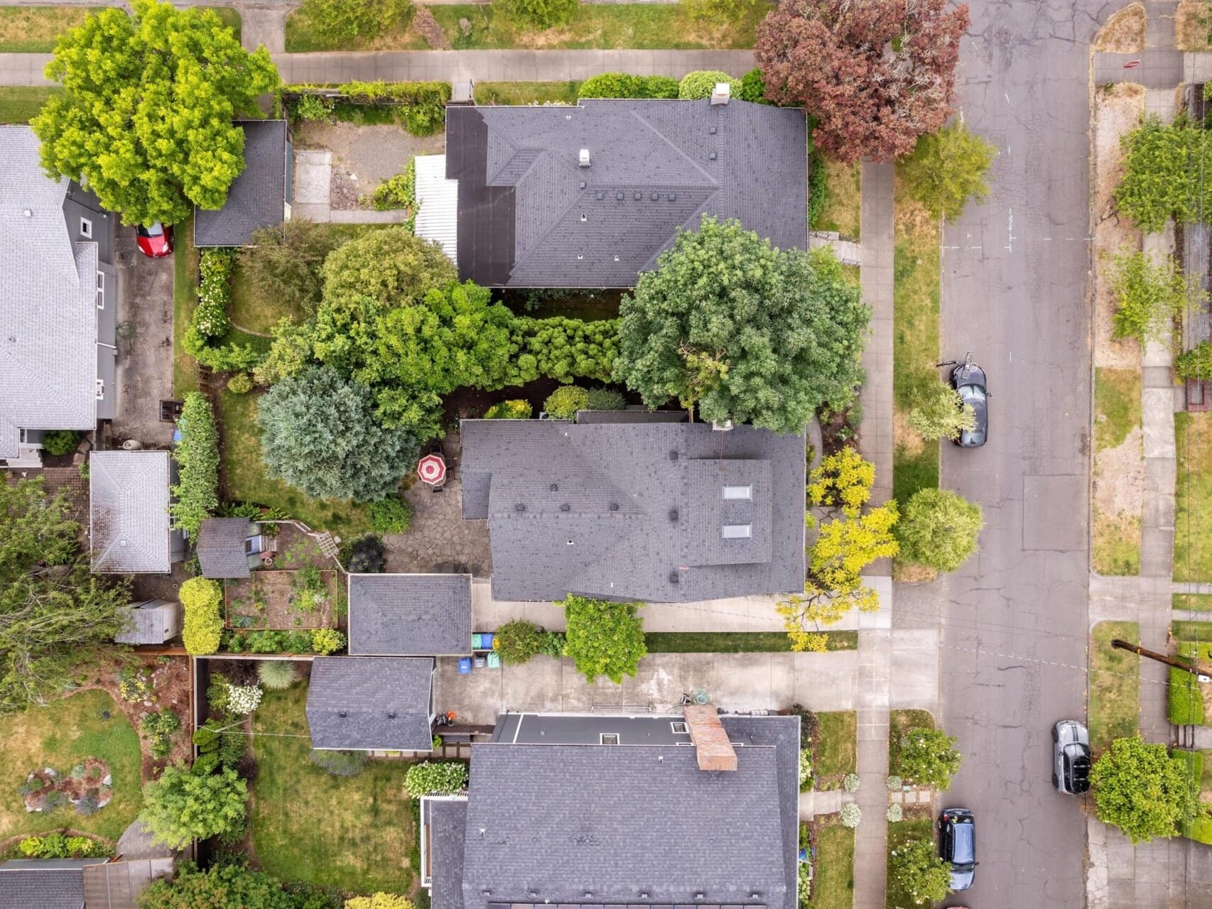 An aerial view of a residential neighborhood featuring several houses with dark roofs, surrounded by green trees and gardens. A street with parked cars lines the area on the right. The scene is captured from directly above.