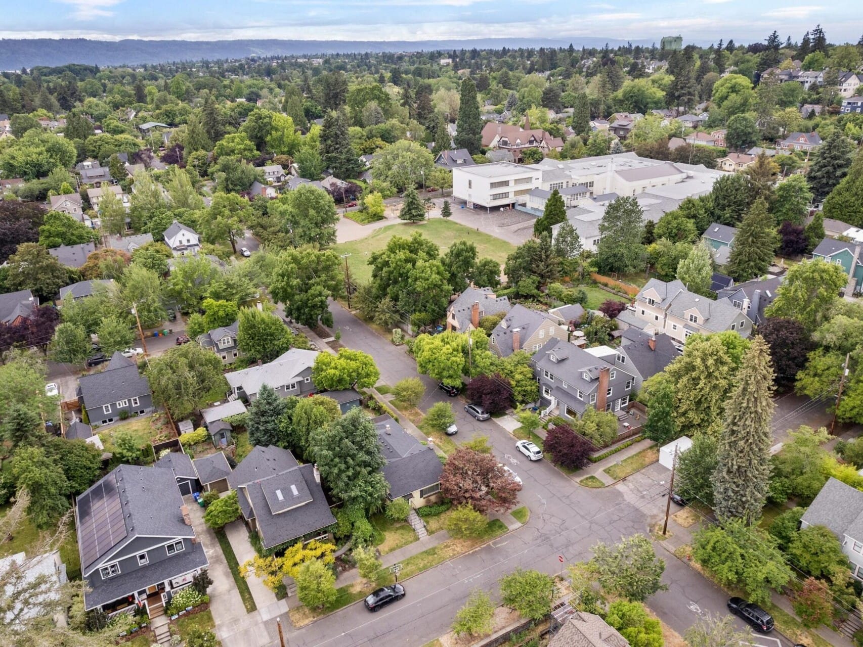 An aerial view of a suburban neighborhood in Portland, Oregon, showcases tree-lined streets and houses nestled amid lush greenery. In the background, a large building rises against a landscape of rolling hills beneath a cloudy sky—a serene snapshot of Portland's real estate charm.