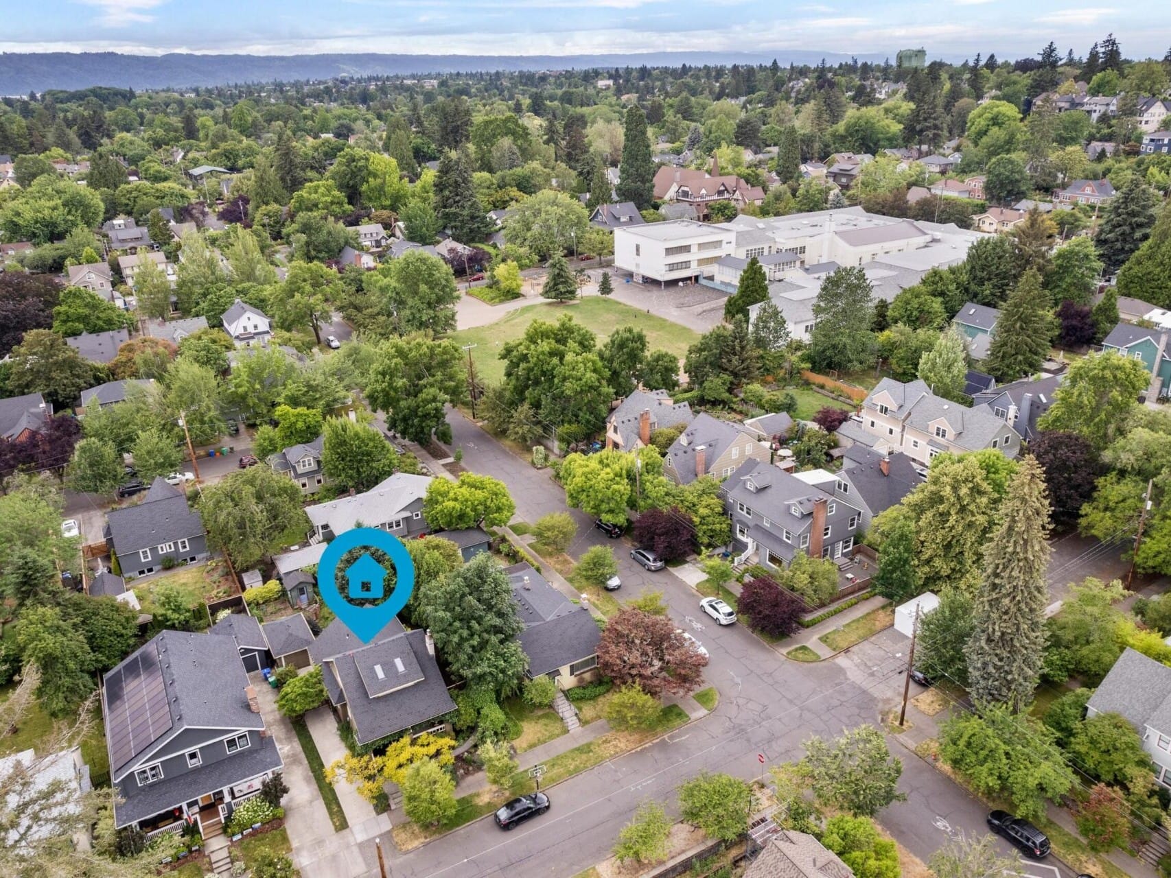 Aerial view of a suburban neighborhood with diverse homes surrounded by trees, highlighting one with a blue location pin. In the background, a large building and distant hills emerge under a partly cloudy sky, showcasing Portland Oregon real estate's charm.