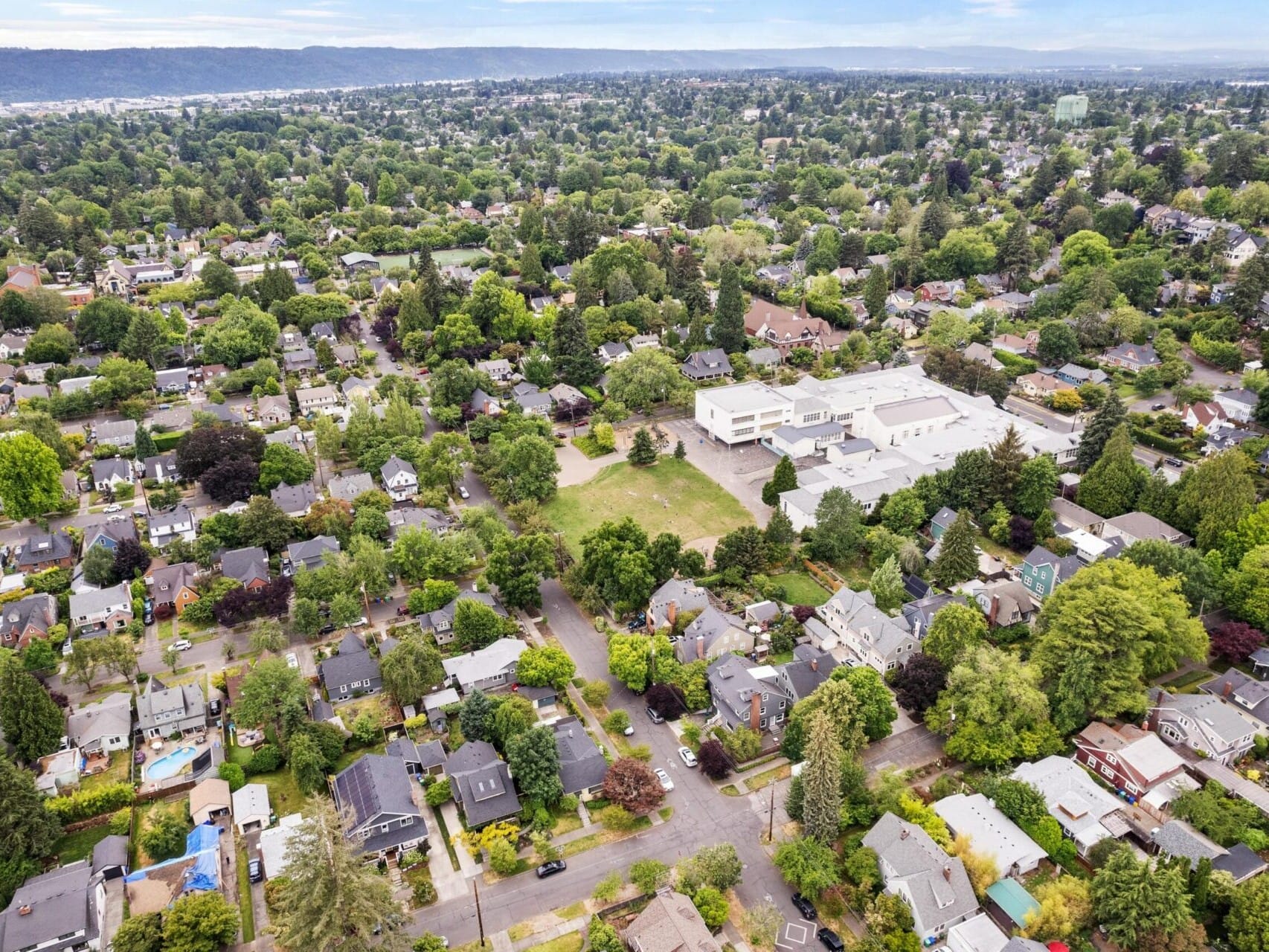 Aerial view of a suburban neighborhood in Portland, Oregon, with numerous houses surrounded by lush greenery. A large building, possibly a school, is centrally located. Roads crisscross through the area, and a distant landscape is visible under a cloudy sky—a snapshot of prime Portland real estate.