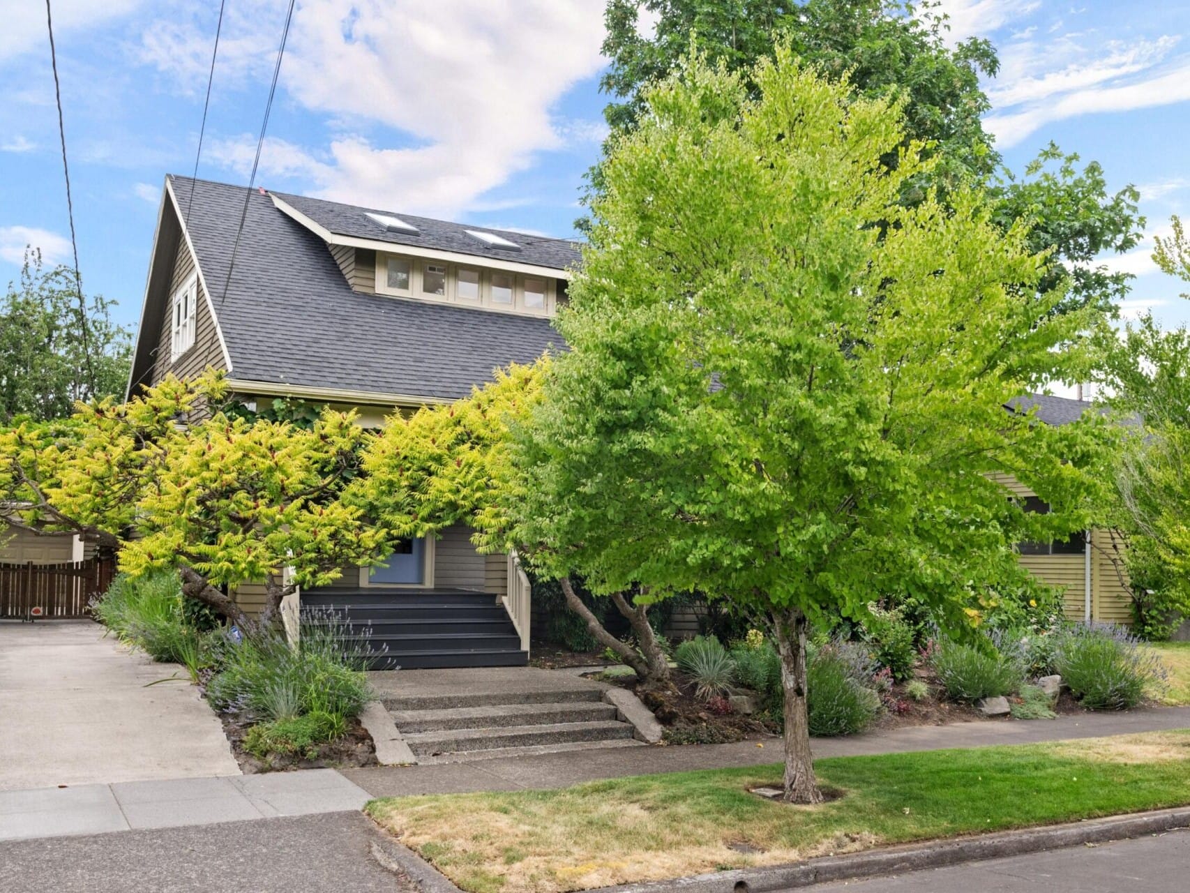 A charming house in Portland with a covered porch and lush greenery. A vine-draped pergola adds character to the front entrance, while trees and shrubs enhance the landscape. With a sloped roof and multiple small windows, it's an enticing find for anyone exploring Portland Oregon real estate.