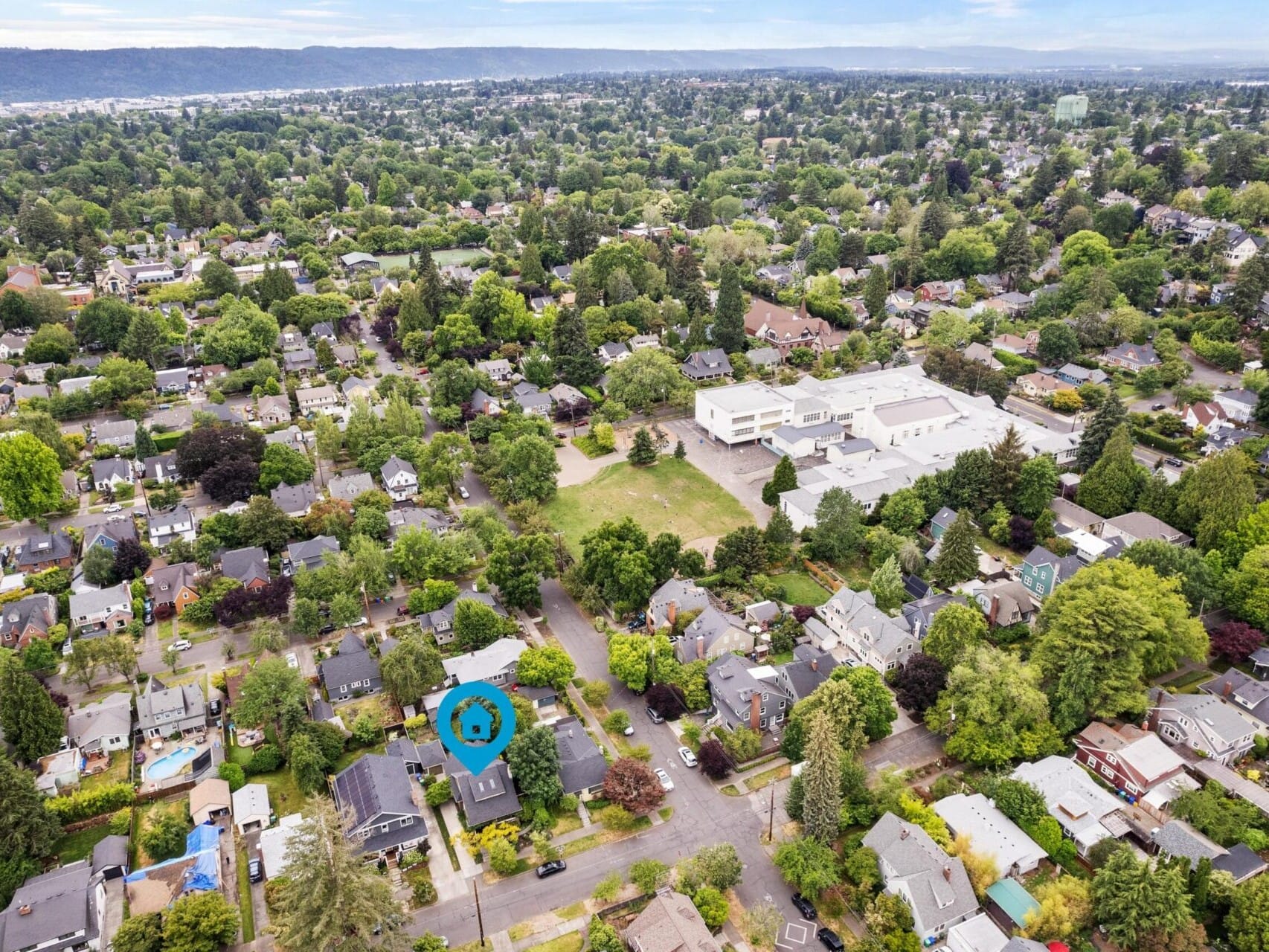 Aerial view of a suburban neighborhood in Portland, Oregon, with numerous houses surrounded by lush green trees. A large white building is visible in the center, marked by a small blue pin highlighting a prime Portland Oregon real estate location. Roads crisscross the area.