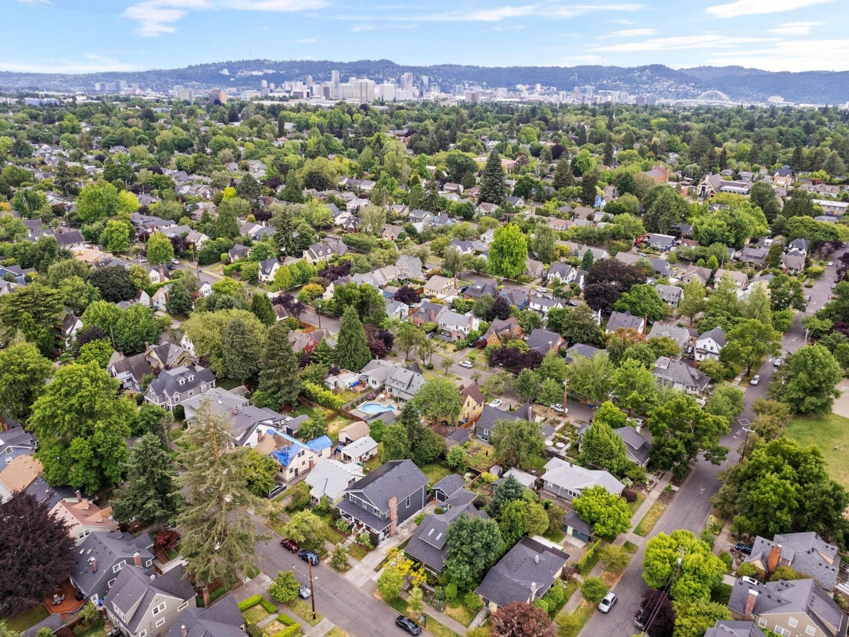 Aerial view of a suburban neighborhood with tree-lined streets and houses. The landscape is lush with greenery, and a distant city skyline is visible under a blue sky, showcasing the charm of Portland Oregon real estate.