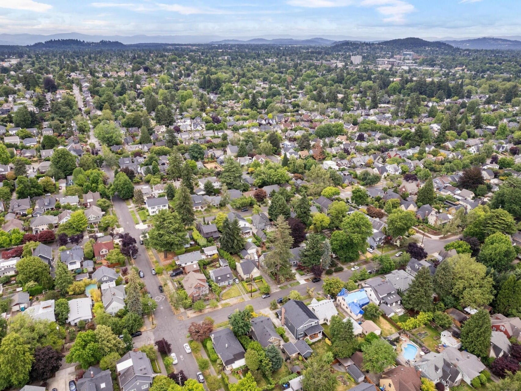 Aerial view of a suburban neighborhood in Portland, Oregon, with tree-lined streets and houses. The area is lush with greenery, ideal for exploring Portland real estate opportunities, as the landscape extends to distant hills under a partly cloudy sky.