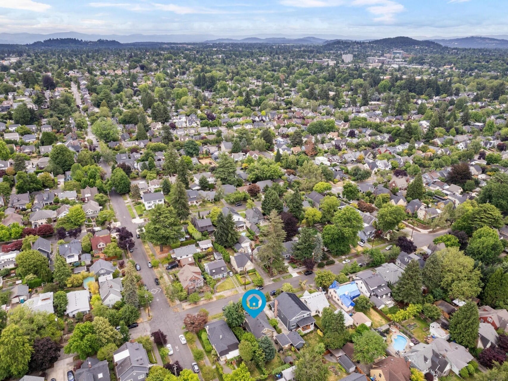 Aerial view of a suburban neighborhood with numerous houses surrounded by lush green trees. Streets intersect at right angles, and a blue marker highlights a specific house, perfect for anyone exploring Portland Oregon real estate. The horizon shows distant hills under a partly cloudy sky.