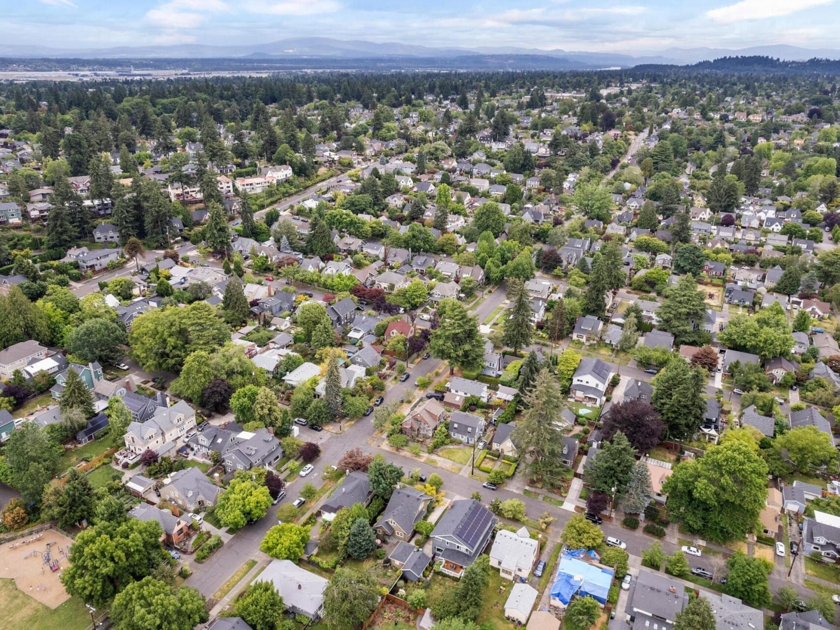 Aerial view of a suburban neighborhood with rows of houses surrounded by lush trees. The landscape, navigated by roads intersecting the area and distant hills under a partly cloudy sky, reflects the charm Portland Oregon Real Estate offers.