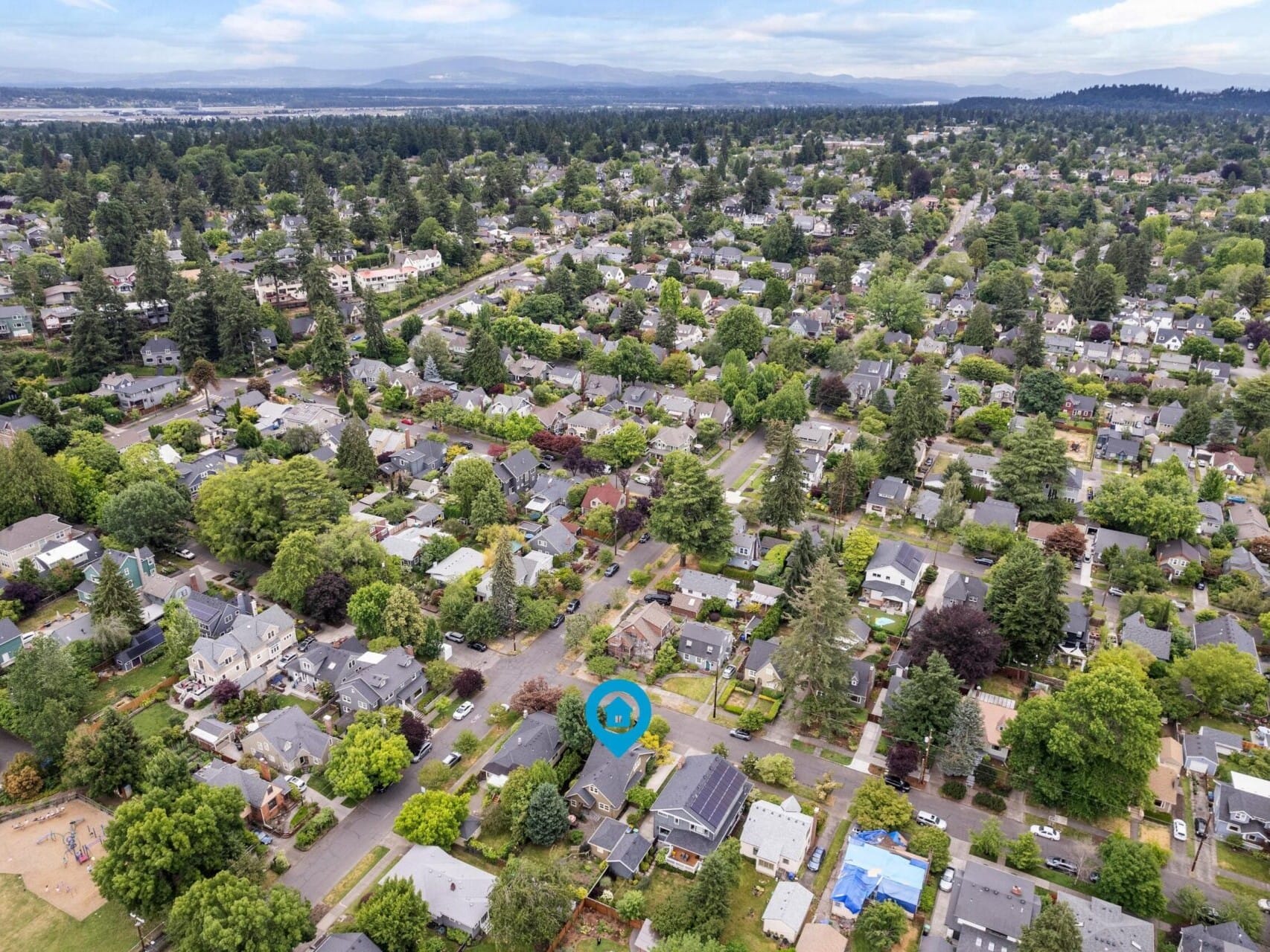 Aerial view of a suburban neighborhood with houses surrounded by lush greenery in the Portland Oregon real estate market. Streets form a grid pattern, marked by a blue circular pin on one house. In the distance, hills and a body of water are visible under a cloudy sky.