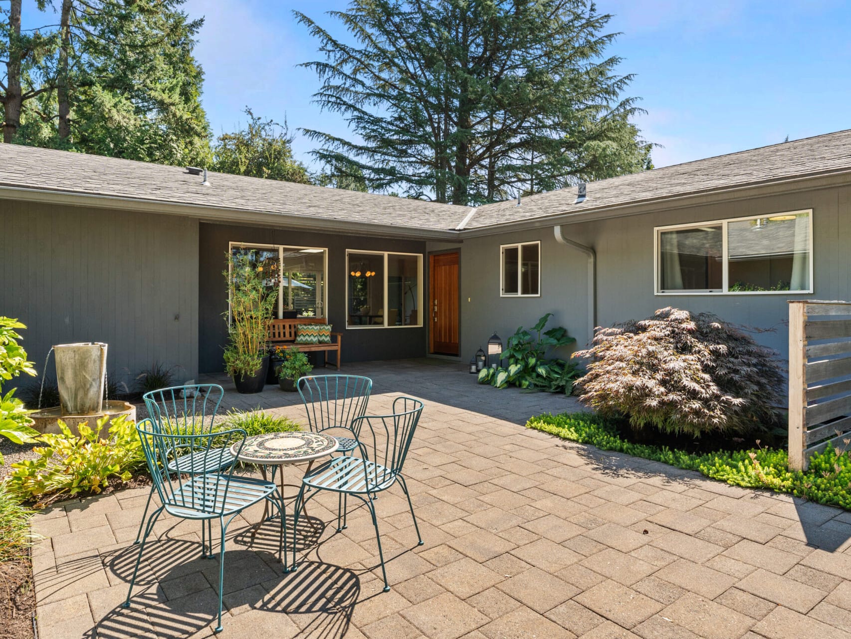 A patio with metal chairs and a table set on a paved area, surrounded by green plants. A gray house with large windows and a wooden door is in the background. Tall trees are visible beyond the house.