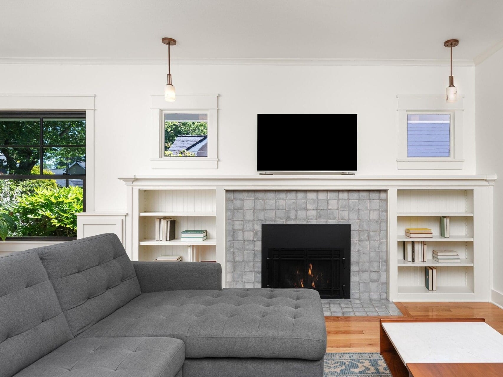 A cozy living room with a gray sectional sofa facing a fireplace. The fireplace features a tile surround and has a black TV mounted above it. Built-in shelves flank the fireplace, and windows on each side allow natural light. A small table is in front.