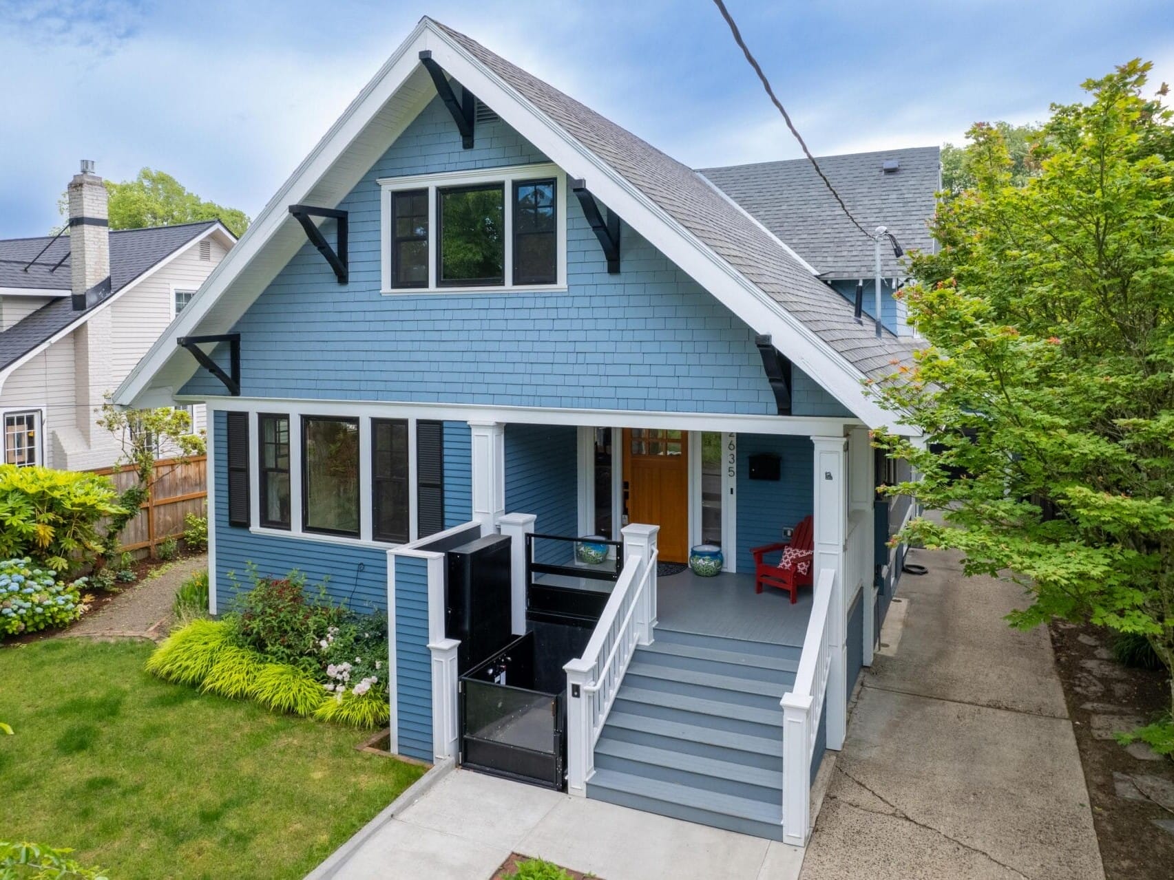 A large blue and white two-story house with a gabled roof, surrounded by greenery. It has a front porch with steps leading up, featuring two red chairs. A concrete driveway is to the right, and neighboring houses are visible.