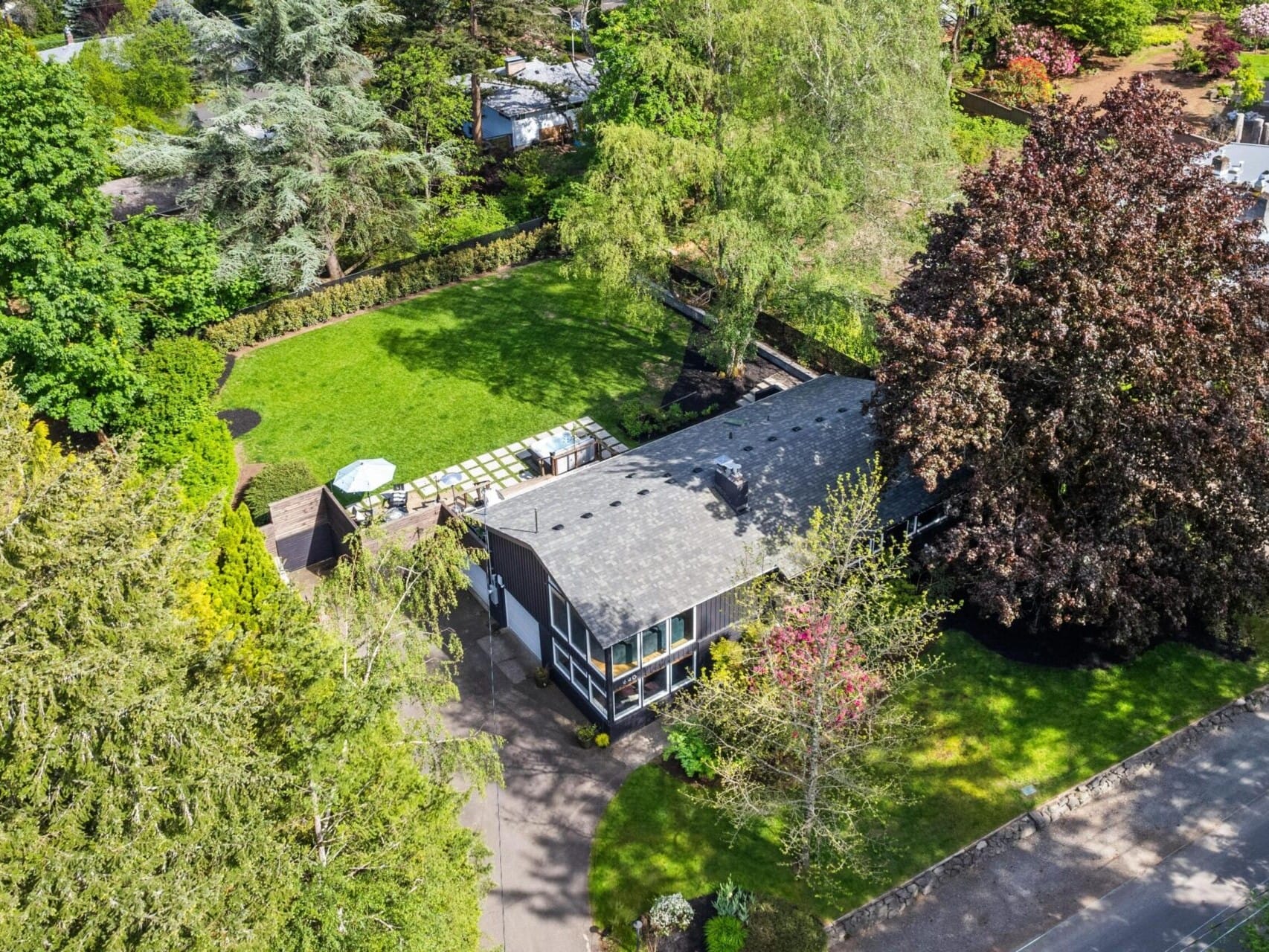 Aerial view of a house surrounded by lush greenery and trees. The house has a gray roof and a spacious lawn. A greenhouse is situated nearby, and a road is visible at the bottom of the image.