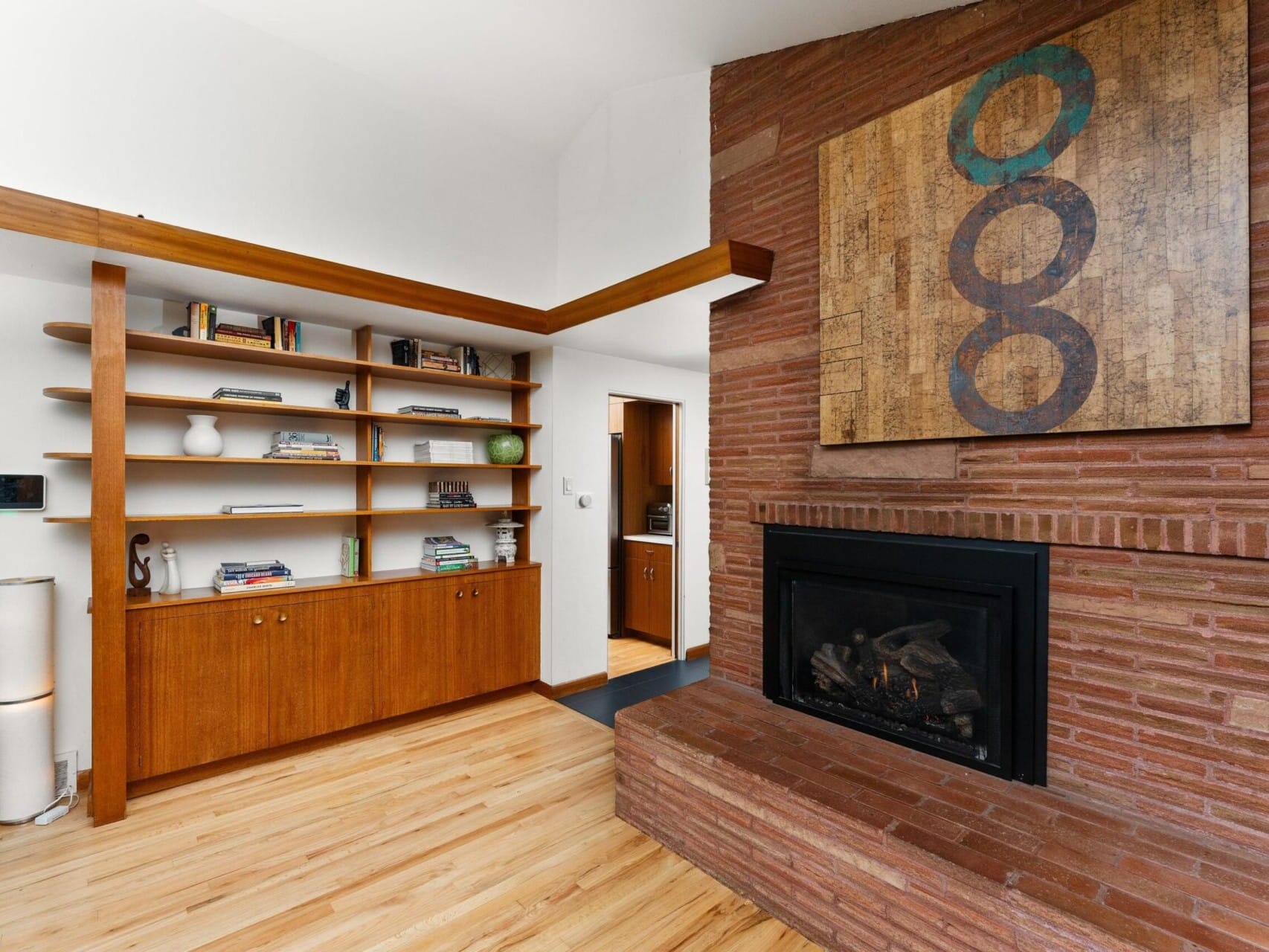 A modern living room features a brick fireplace with a large abstract painting above it. To the left, theres a wooden bookshelf with books and decorative items. The room has a light wood floor and a potted plant in the corner.