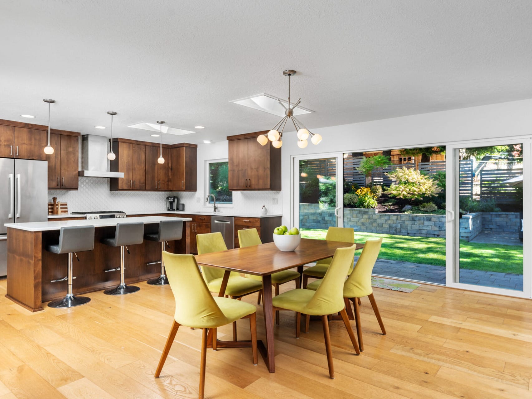 A modern kitchen and dining area with wooden flooring, brown cabinets, and stainless steel appliances. A rectangular table with six yellow chairs is centered under a contemporary light fixture. Sliding glass doors open to a landscaped backyard.