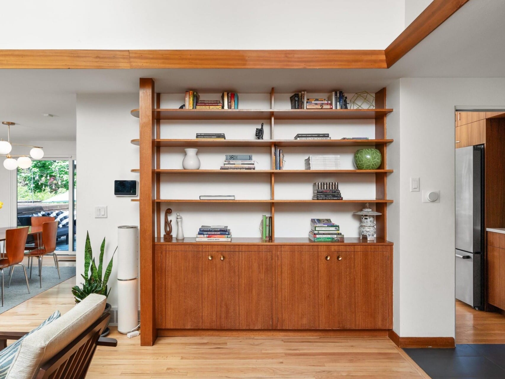 A modern living space featuring a built-in wooden bookshelf with shelves full of books and decorative items. To the left, a glimpse of the dining area with a table, chairs, and a vase of yellow flowers. The room is bright with ample natural light.