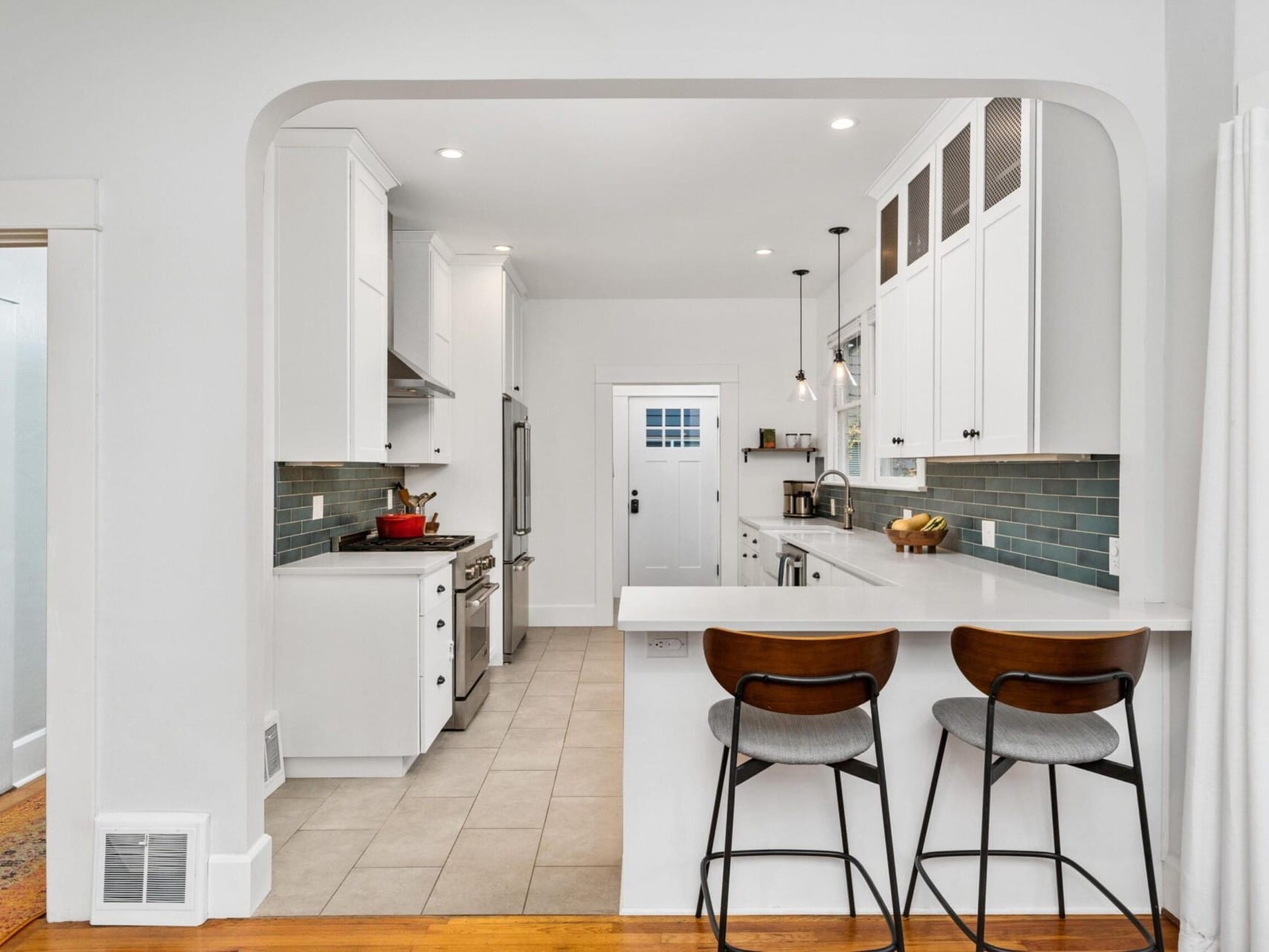 A modern kitchen with tile flooring, white cabinetry, and a gray-tiled backsplash. Features a white countertop bar with two wooden stools, a gas stove with a red pot, and a fruit bowl. A door at the end leads outside. White walls and ceiling lights.