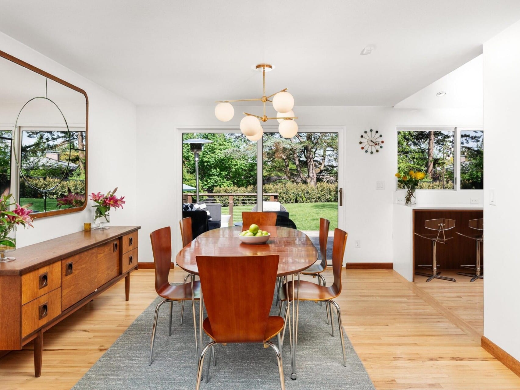 A modern dining room with a wooden table and six chairs on a gray rug. A wooden sideboard with flowers and a mirror is on the left. A spherical light fixture hangs above. A window and sliding door offer views of a garden. Two stools are by a counter.