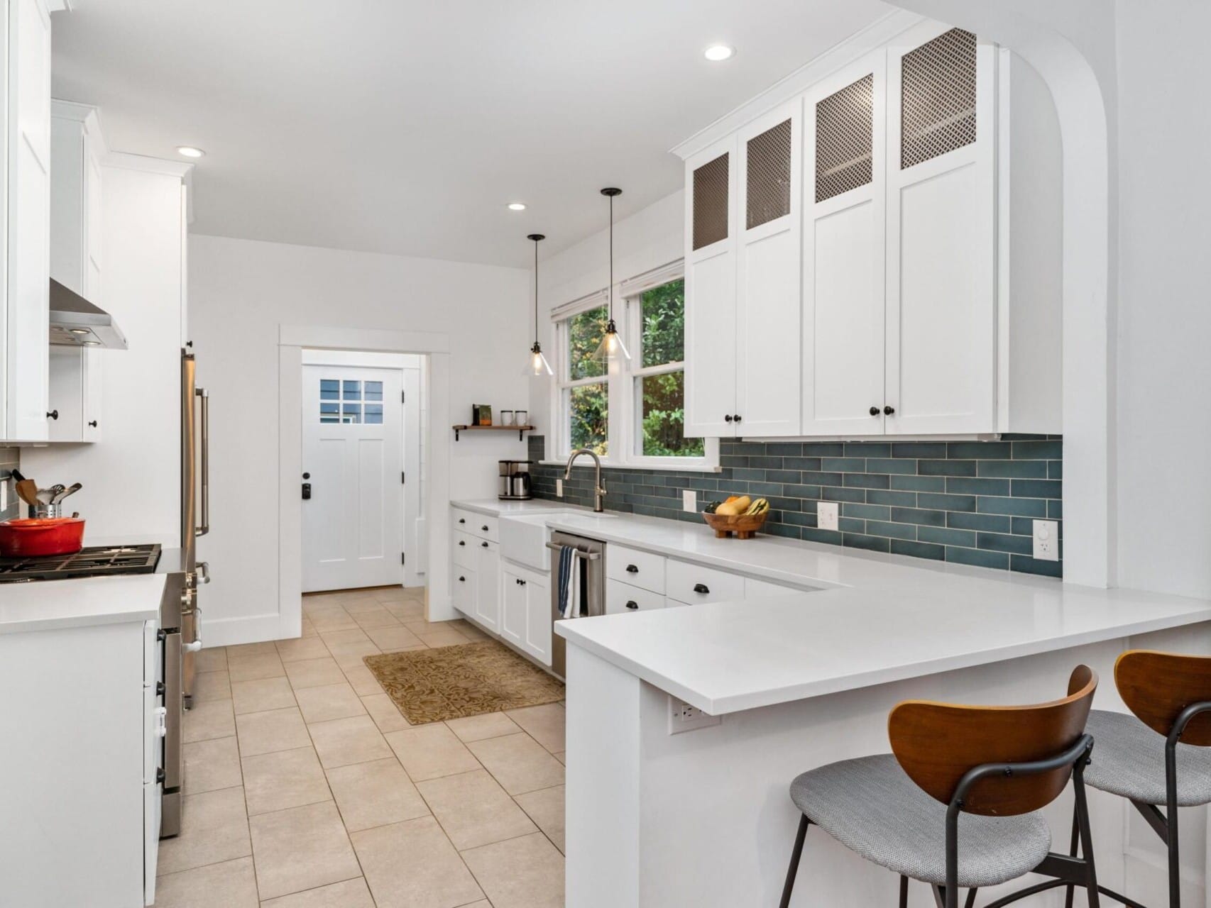 A modern kitchen with white cabinets and blue tile backsplash. It features stainless steel appliances, a double sink, and a bar with two wooden stools. Large windows provide natural light, and a door leads to the outside. A red pot sits on the stove.