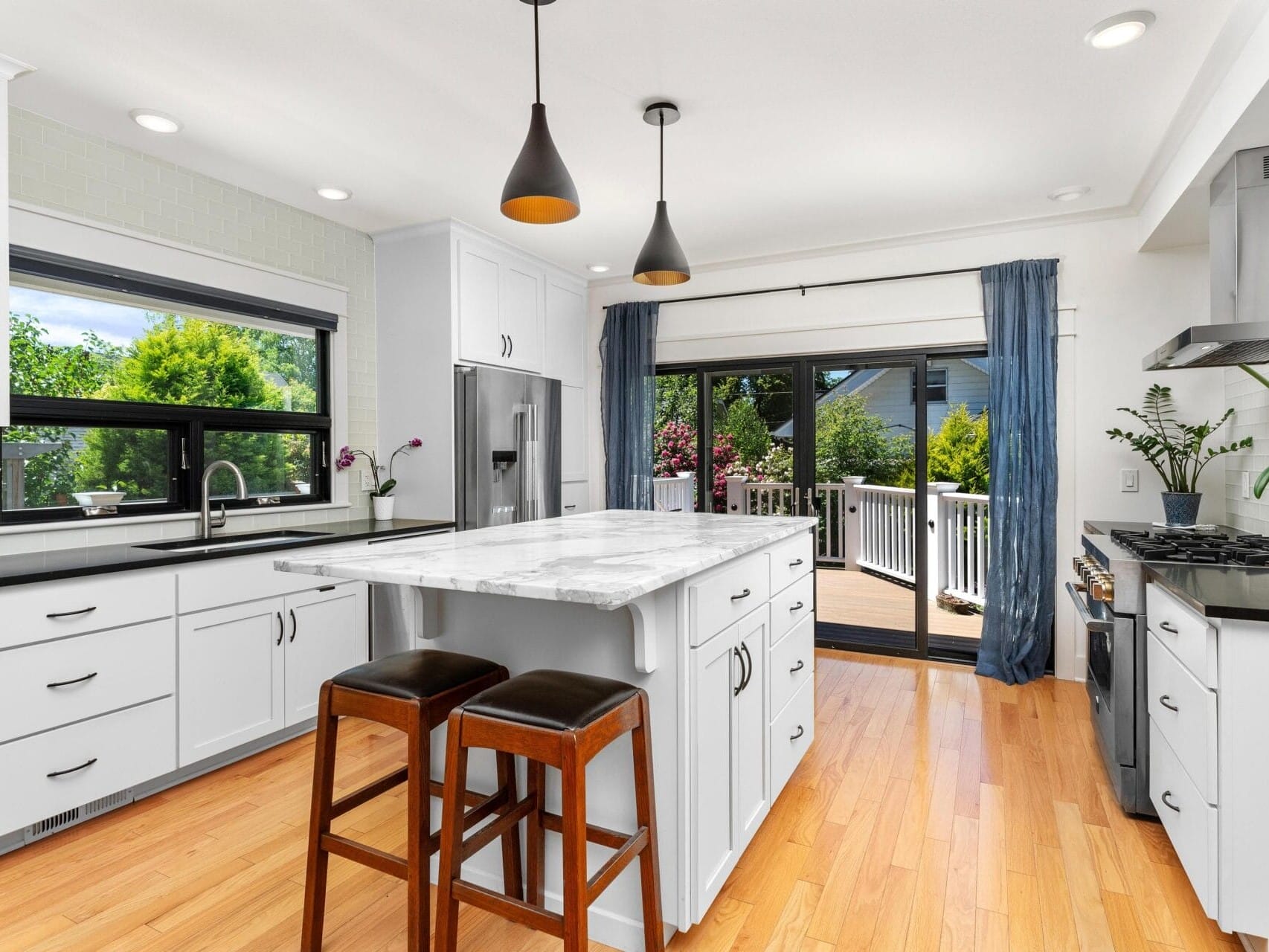 A modern kitchen with white cabinetry, a marble island, and two wooden stools. There are hanging pendant lights, a sink under a window, a gas stove, and glass doors leading to a deck with garden views. The floor is light wood.