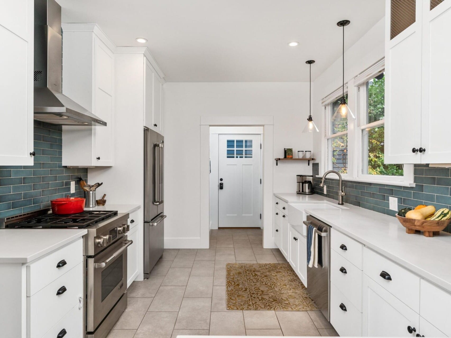 A modern kitchen with white cabinets, stainless steel appliances, and a blue tile backsplash. A red pot sits on the stove, and pendant lights hang above the white countertop. A door with a window is at the end of the room.