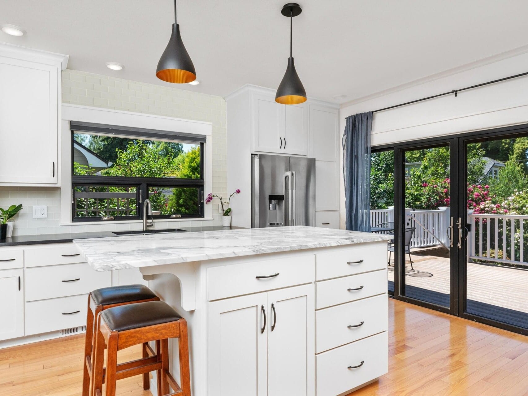 Modern kitchen with white cabinets, a marble island, and two brown stools. Black pendant lights hang overhead. Large windows and glass doors open to a deck, allowing sunlight in. Stainless steel appliances and plants add a fresh touch.