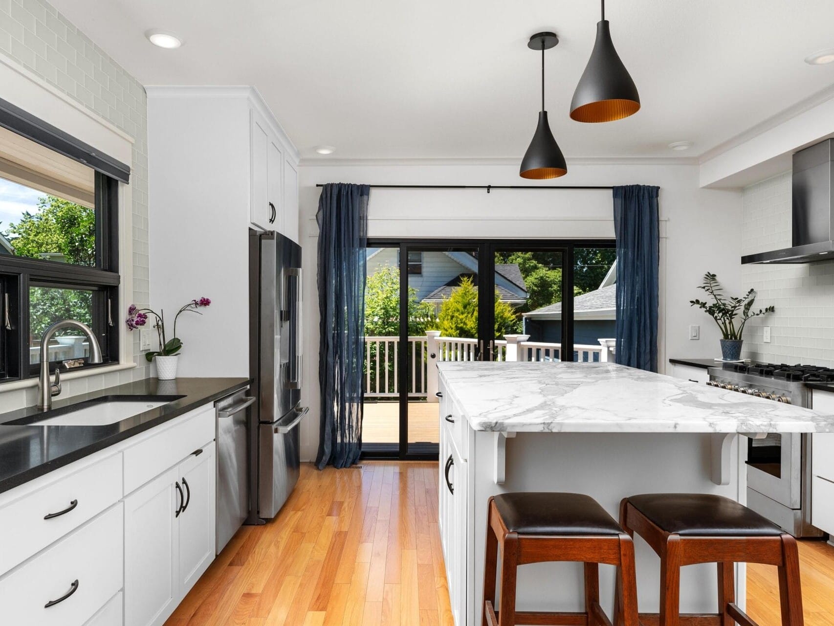 Modern kitchen with white cabinets, black countertops, and a marble kitchen island with two brown stools. Large windows and sliding glass doors open to a deck with greenery visible outside. Pendant lights hang above the island.