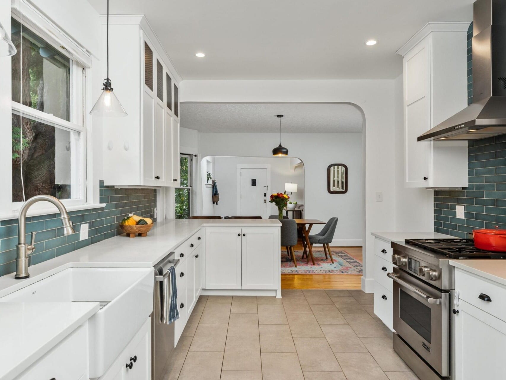 A modern kitchen with white cabinets, a stainless steel stove, and a farmhouse sink. Blue tiles form the backsplash. A red pot sits on the stove. In the background, a dining table with chairs and a light fixture are visible through an archway.