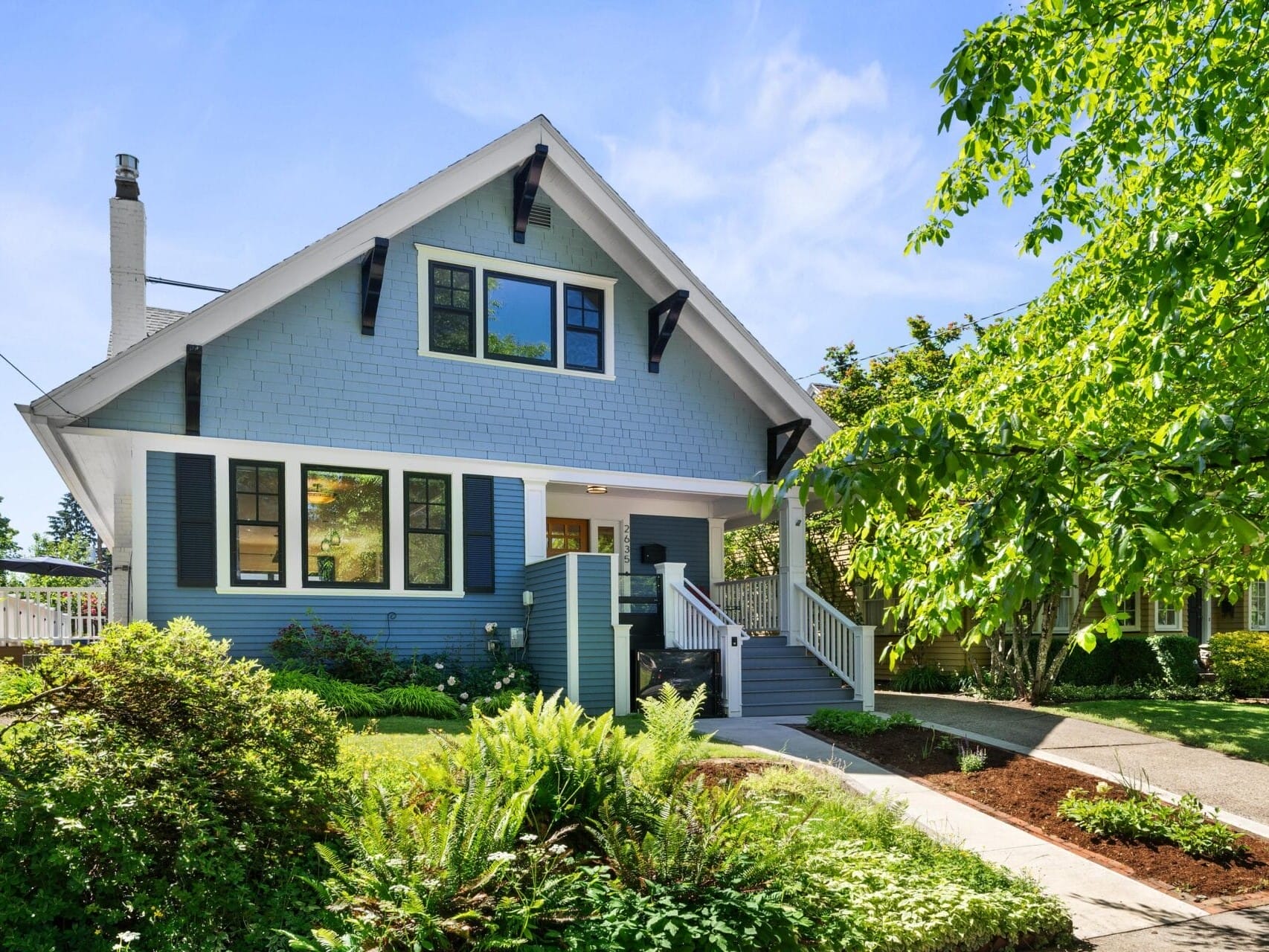 A charming blue house with a steep gabled roof, black shutters, and a white front porch. It is surrounded by lush green landscaping, including shrubs, trees, and a neatly maintained lawn under a clear blue sky.