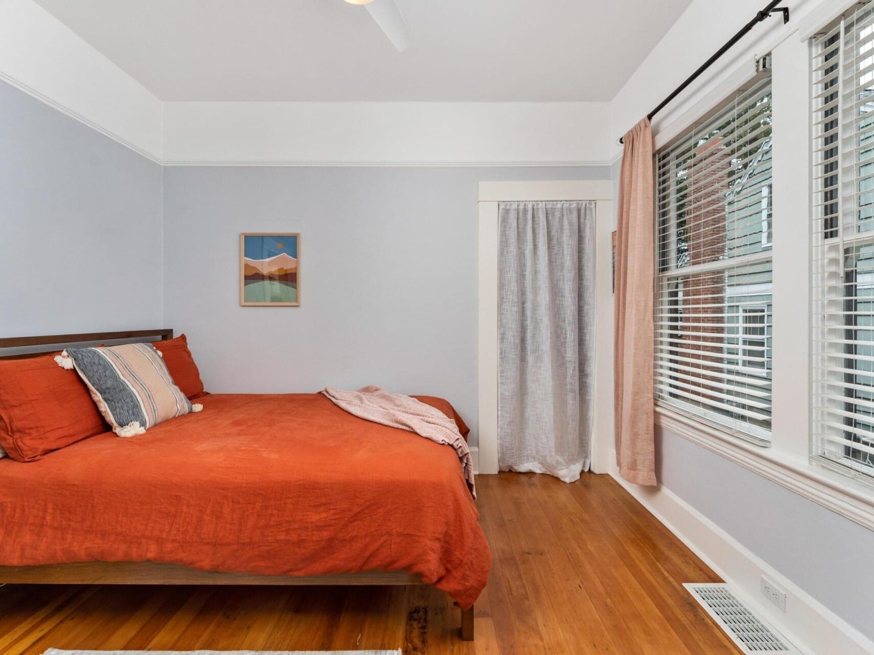 A bedroom with a wooden floor features a bed with orange bedding and pillows. A colorful artwork hangs above the bed. A large window with blinds and pink curtains is on the right. A white door with a gray curtain is near the bed.