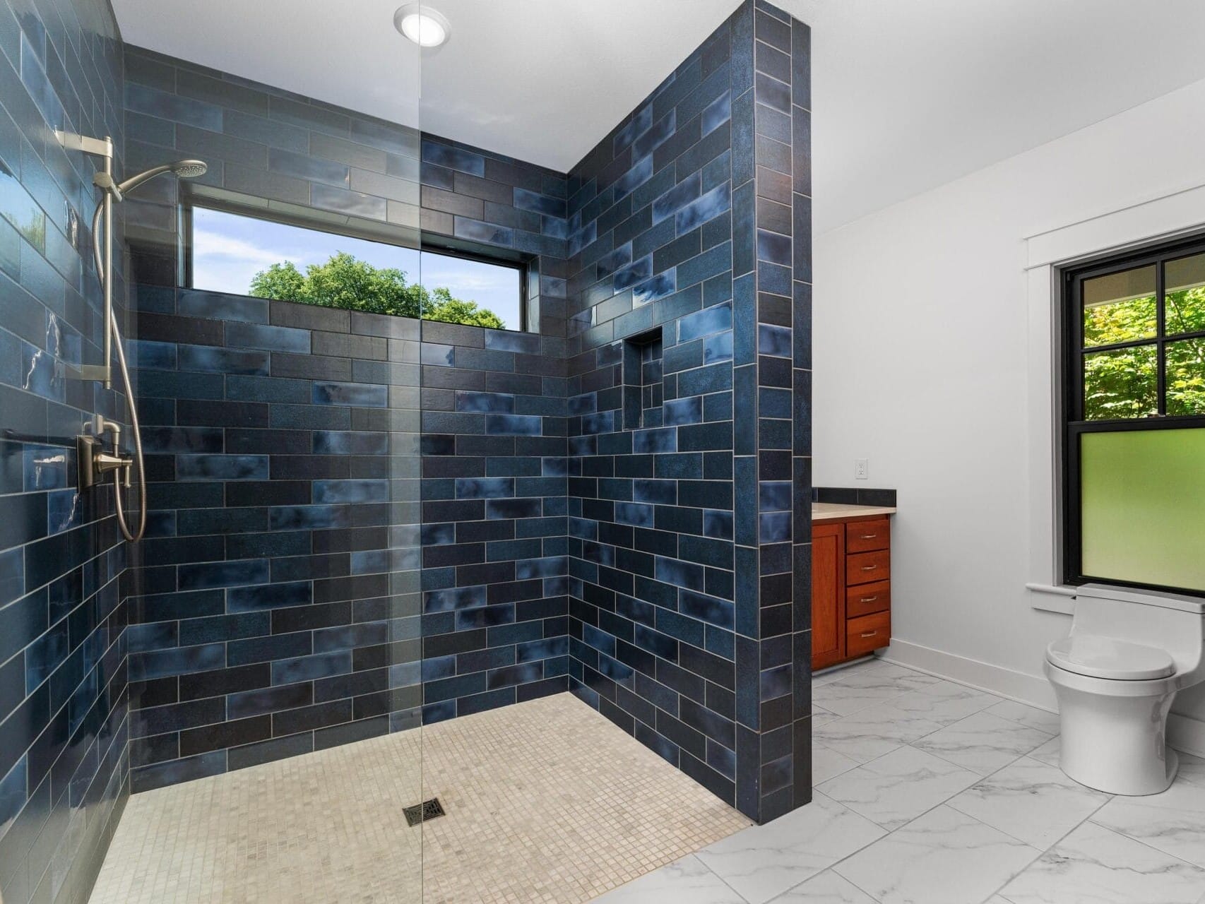 A modern bathroom features a spacious shower with dark blue tile walls and a large window letting in natural light. The floor is tiled with a mosaic pattern, and the vanity is equipped with wooden cabinetry. A white toilet is nearby.