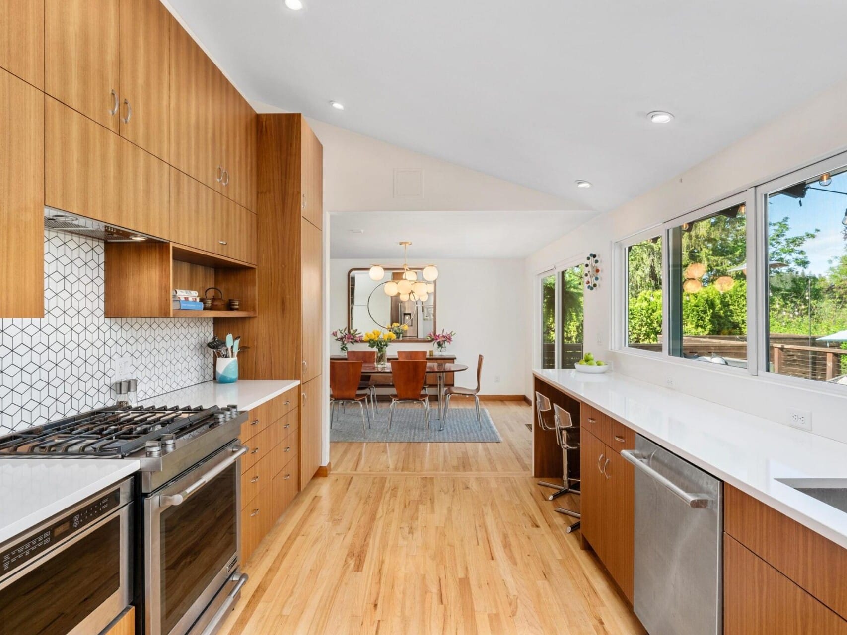 A modern kitchen with wooden cabinets and stainless steel appliances on the left. A spacious dining area with a wooden table and chairs is visible in the background. Large windows on the right provide natural light and a view of greenery outside.