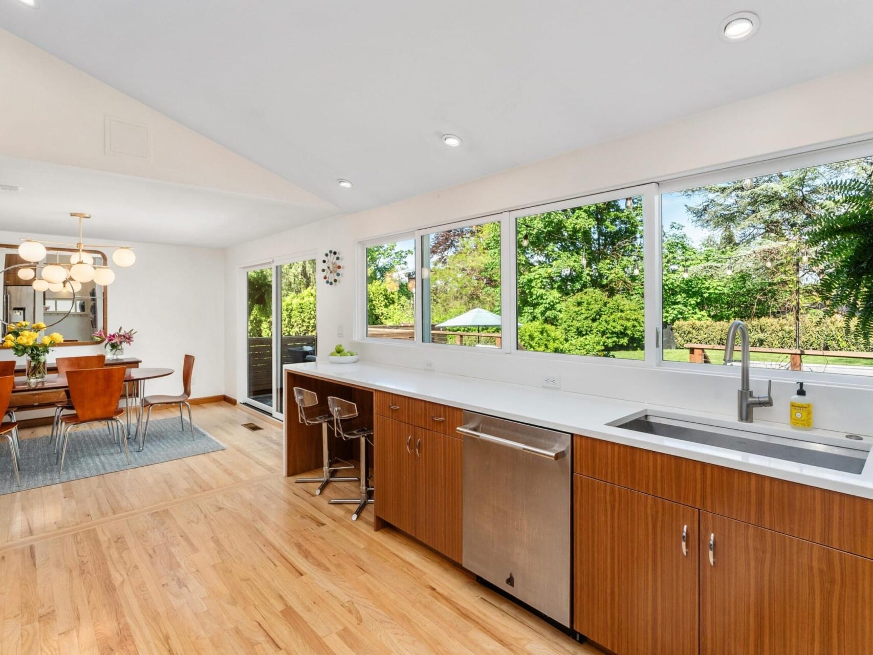 A modern kitchen with wooden cabinets, a white countertop, and stainless steel appliances. Large windows offer views of a lush garden. Adjacent is a dining area with a round table, orange chairs, and a chandelier above. Natural light fills the space.