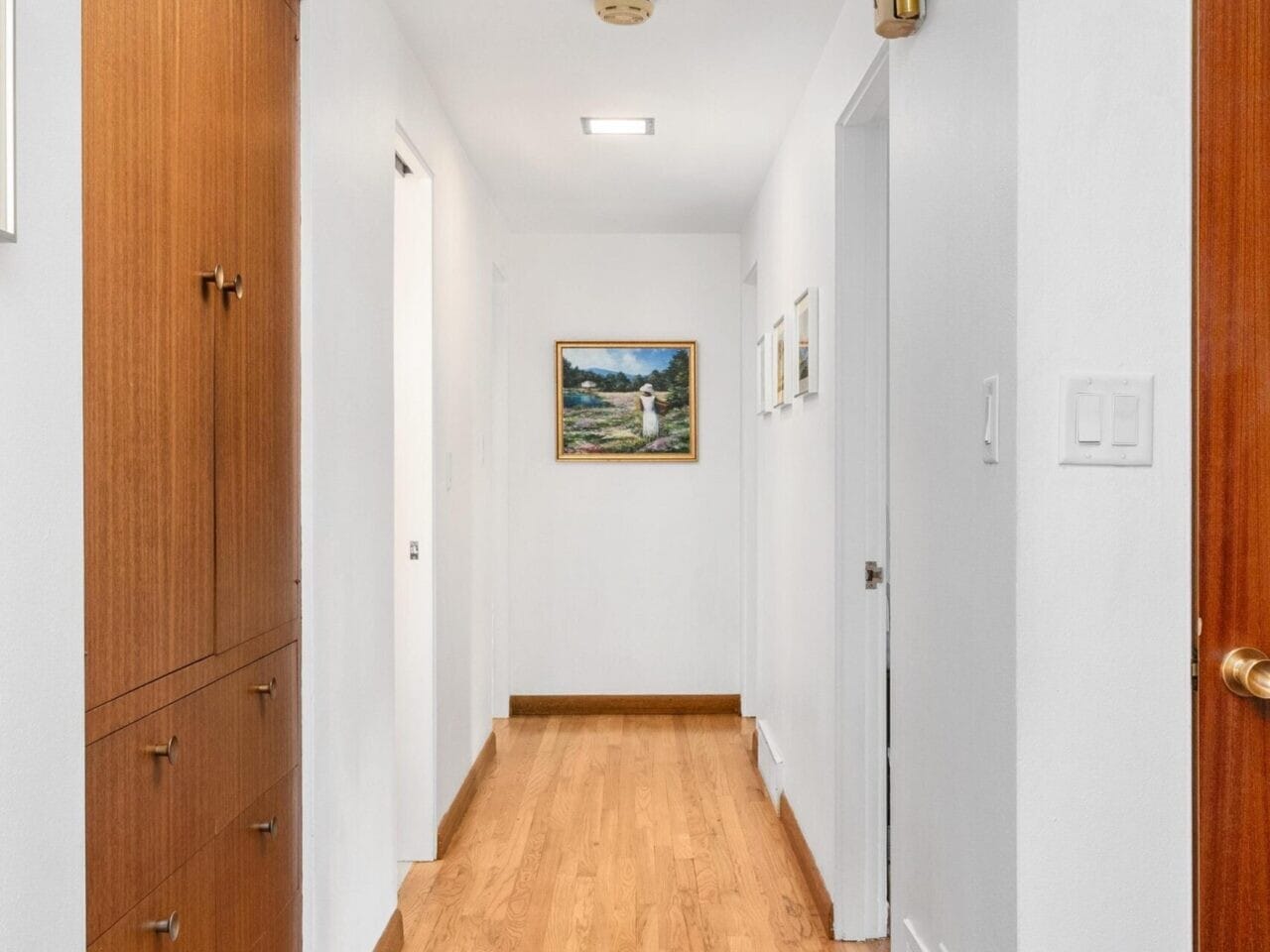 A hallway with wooden flooring and white walls. On the left, there are wooden cabinets, and on the right, a few closed doors. A framed painting of a landscape is hung at the end of the hallway.