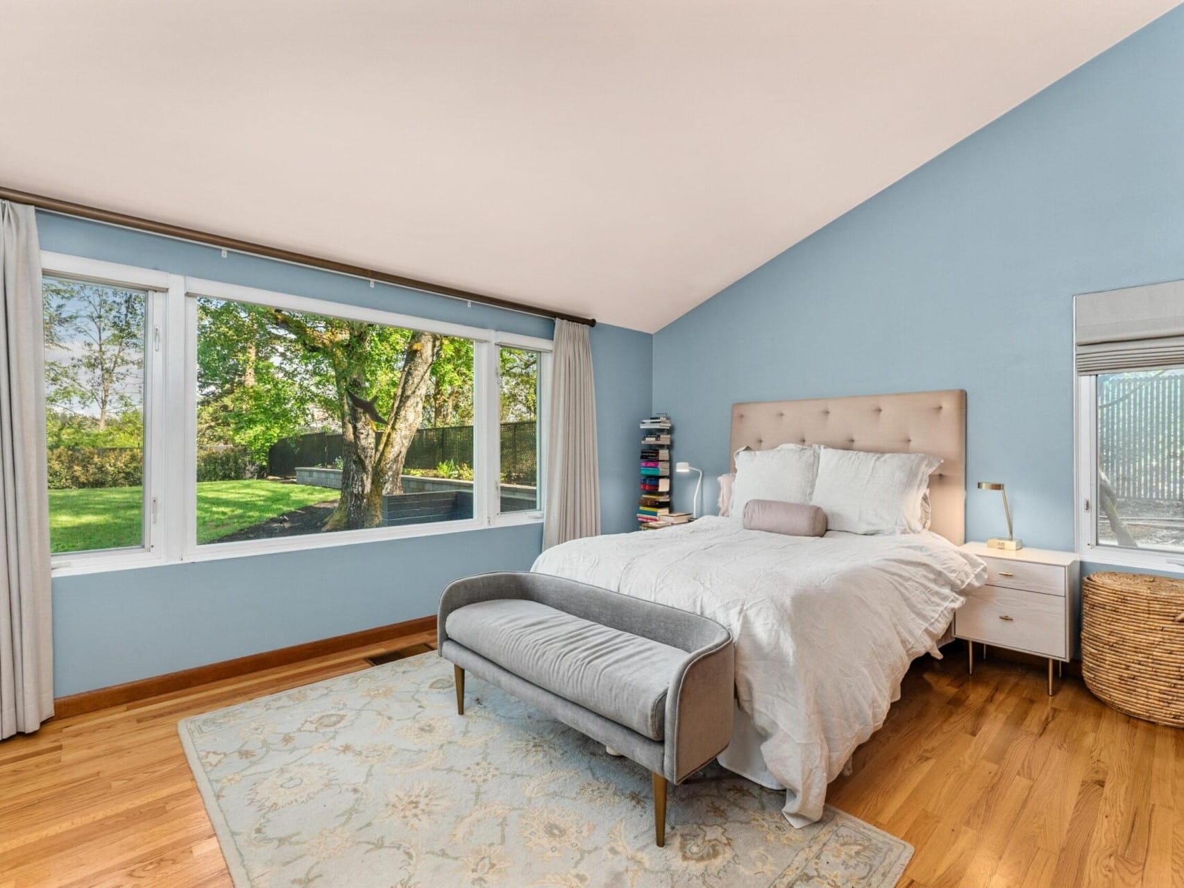 A bedroom with light blue walls and a large window overlooking a garden. It features a bed with a tufted headboard, a gray bench, a nightstand with a lamp, and a rug on a wooden floor. Drapes frame the window, letting in natural light.