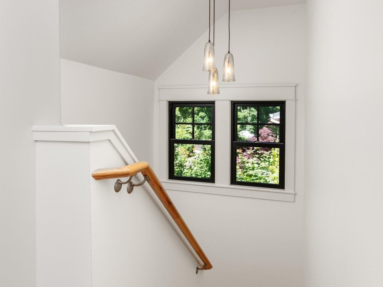 A small staircase with wooden steps and a wooden handrail leads down to a landing. Three pendant lights hang from the ceiling. Two black-framed windows reveal greenery outside, surrounded by white walls.