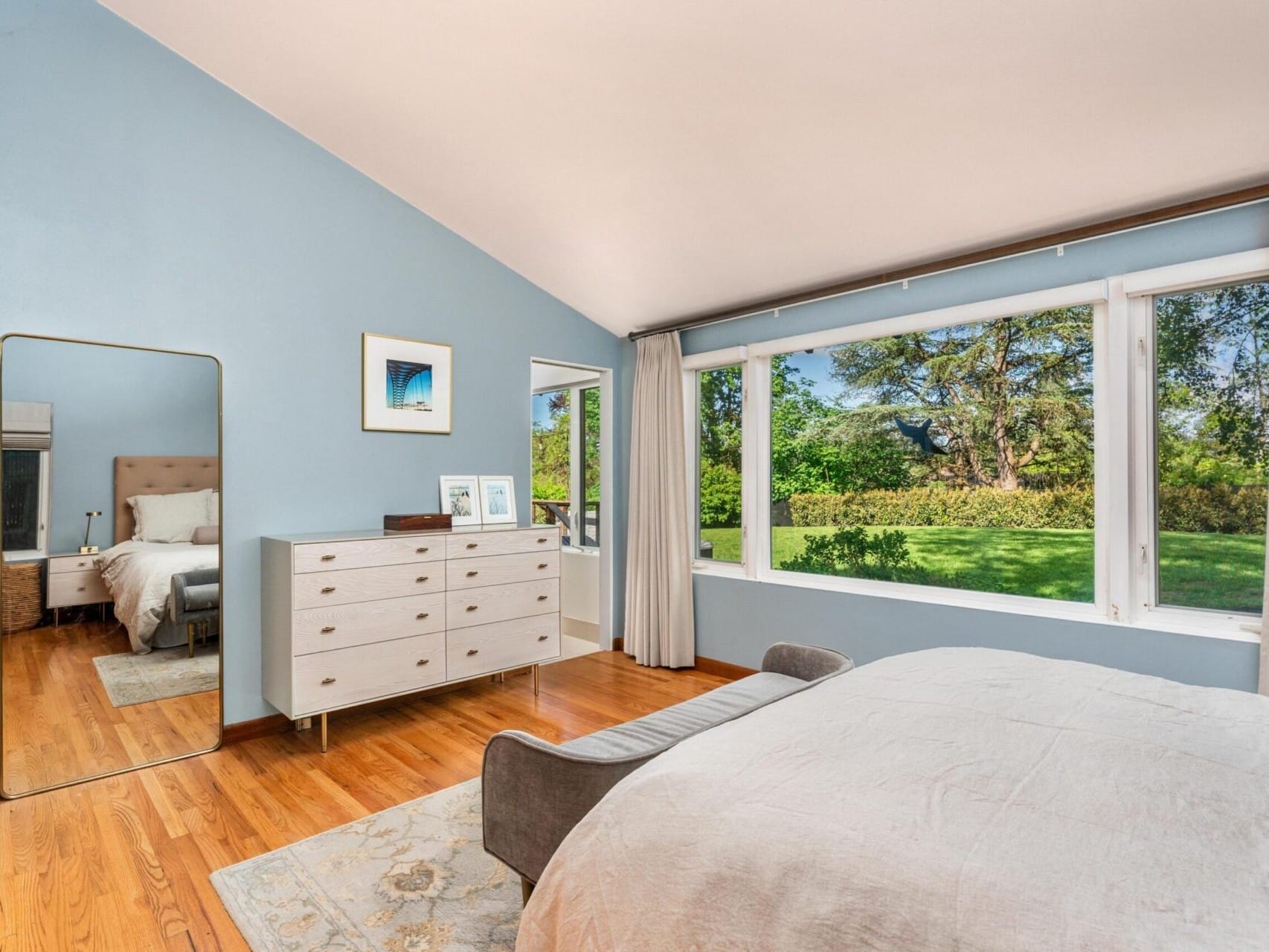 A serene bedroom with light blue walls features a large window overlooking a green backyard. The wooden floor complements a light-colored bed, dresser, and a tall mirror. Neutral curtains frame the window, adding warmth to the space.